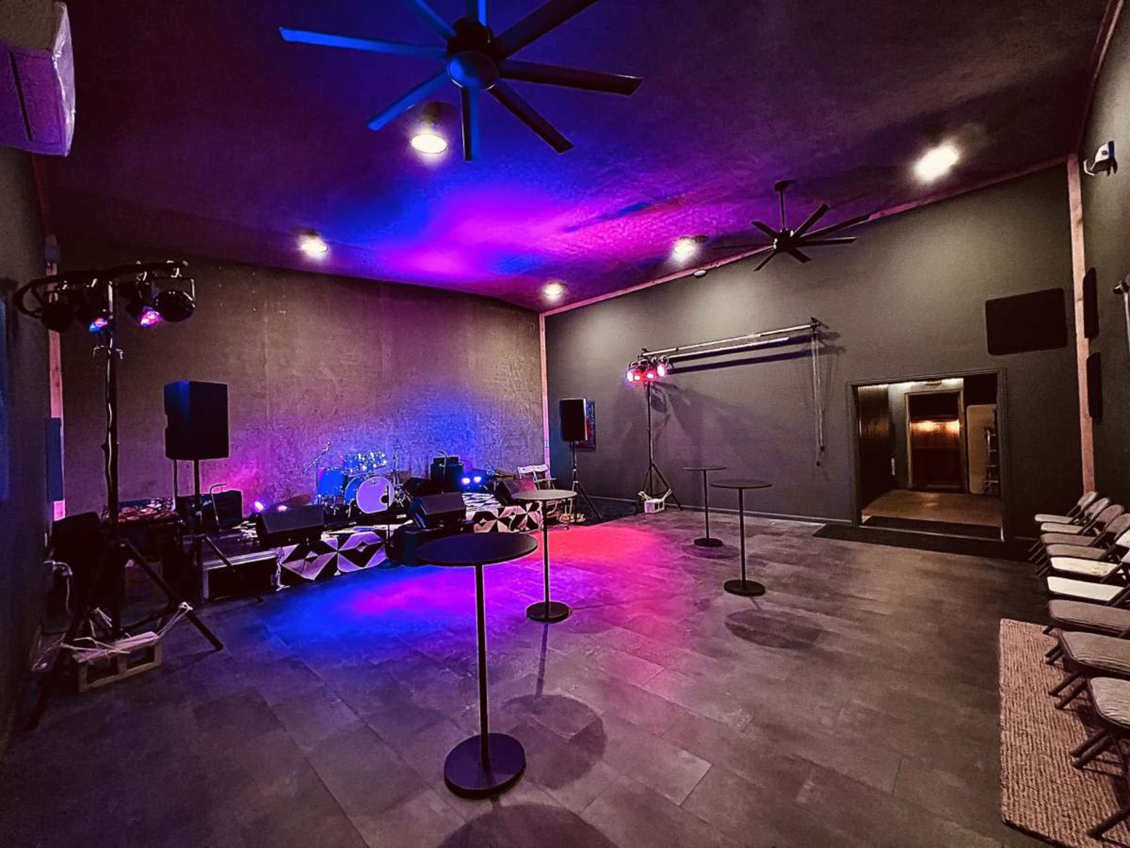 The image shows a dimly lit event space with colorful lighting, featuring music equipment, tables, and chairs arranged for a gathering.