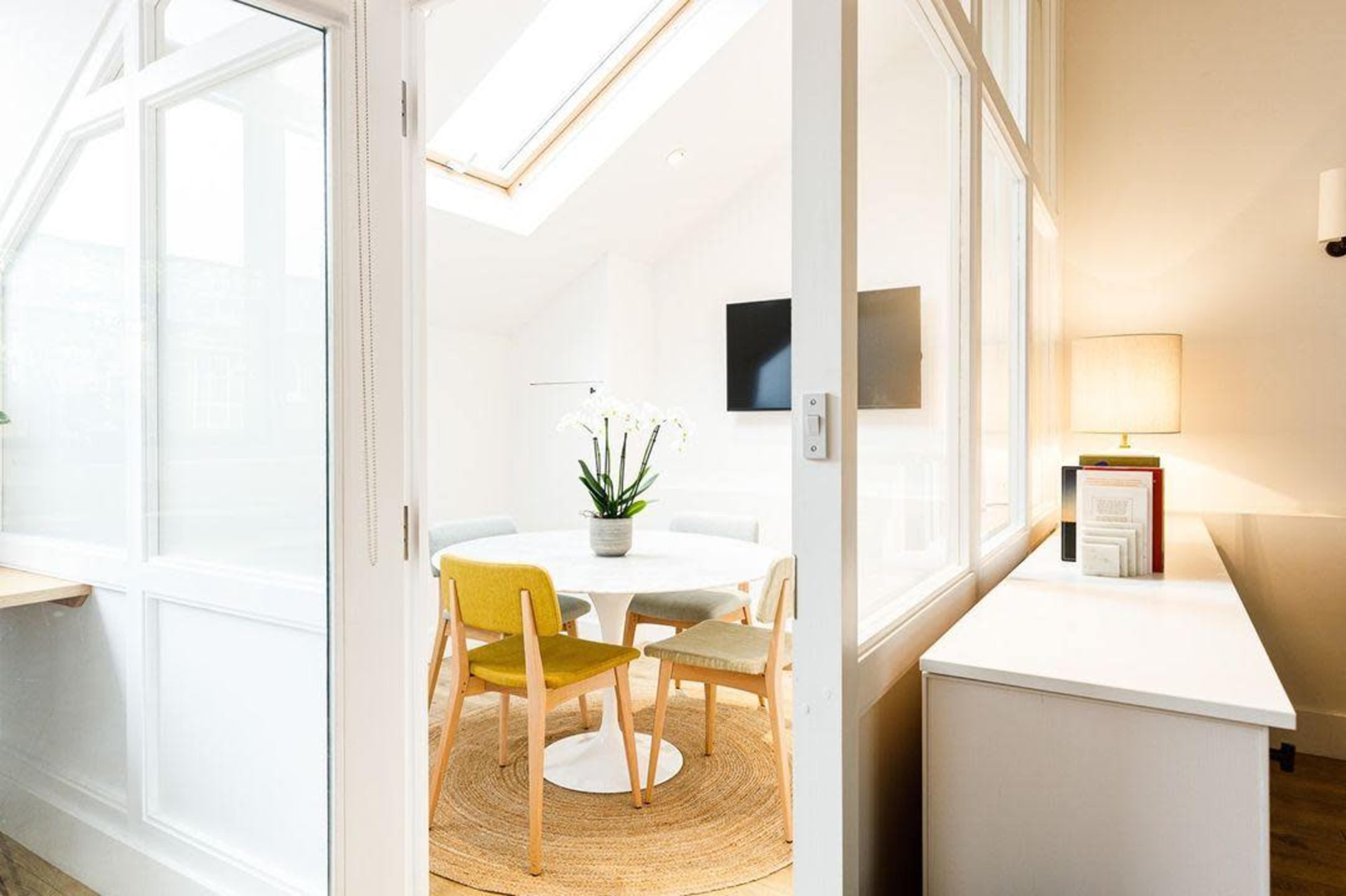 A bright room features a round table surrounded by chairs, with a plant centerpiece and a lamp on a nearby side table.