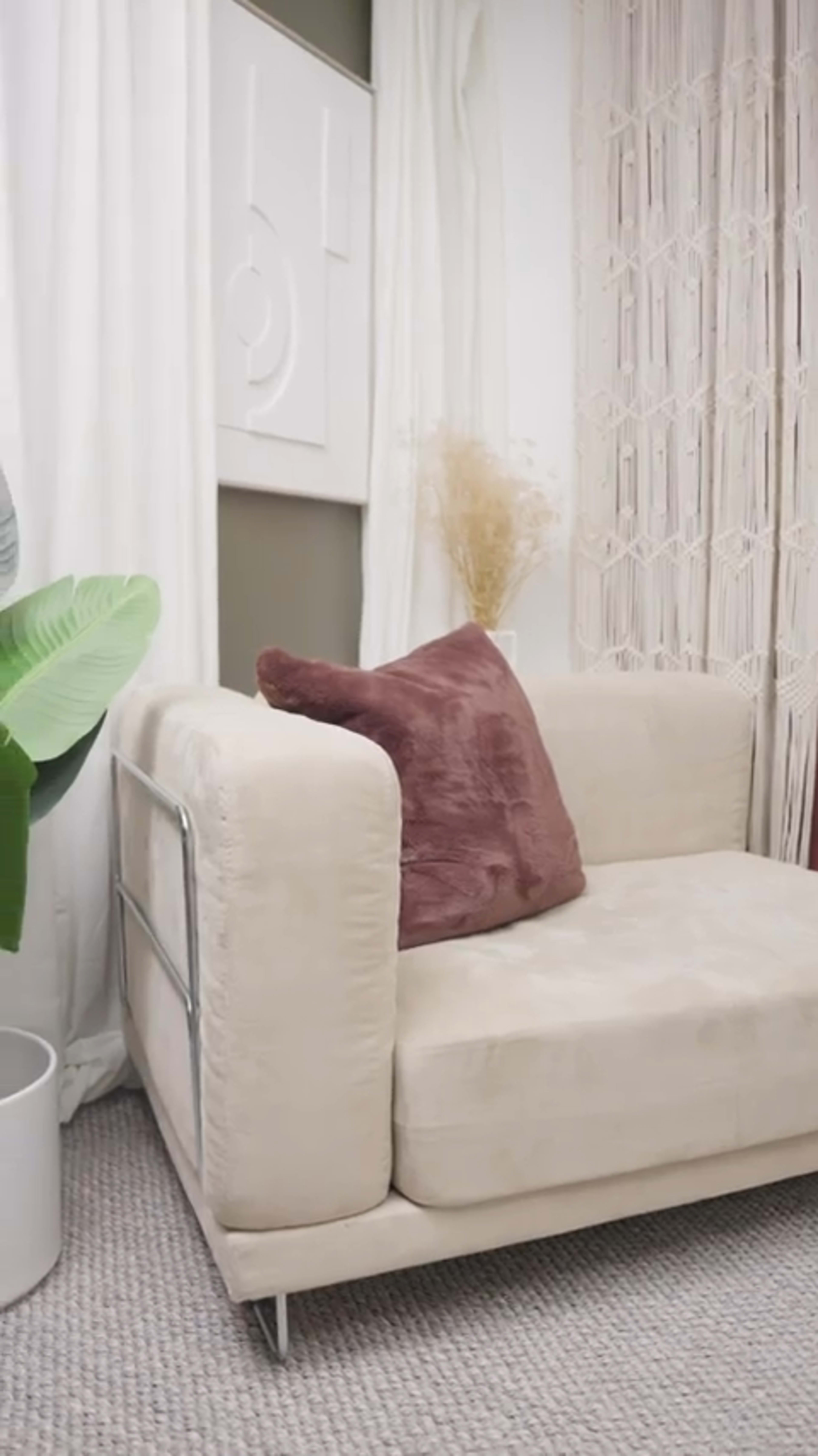 A modern living room corner featuring a beige couch with a brown pillow, decorative plants, and textured curtains.