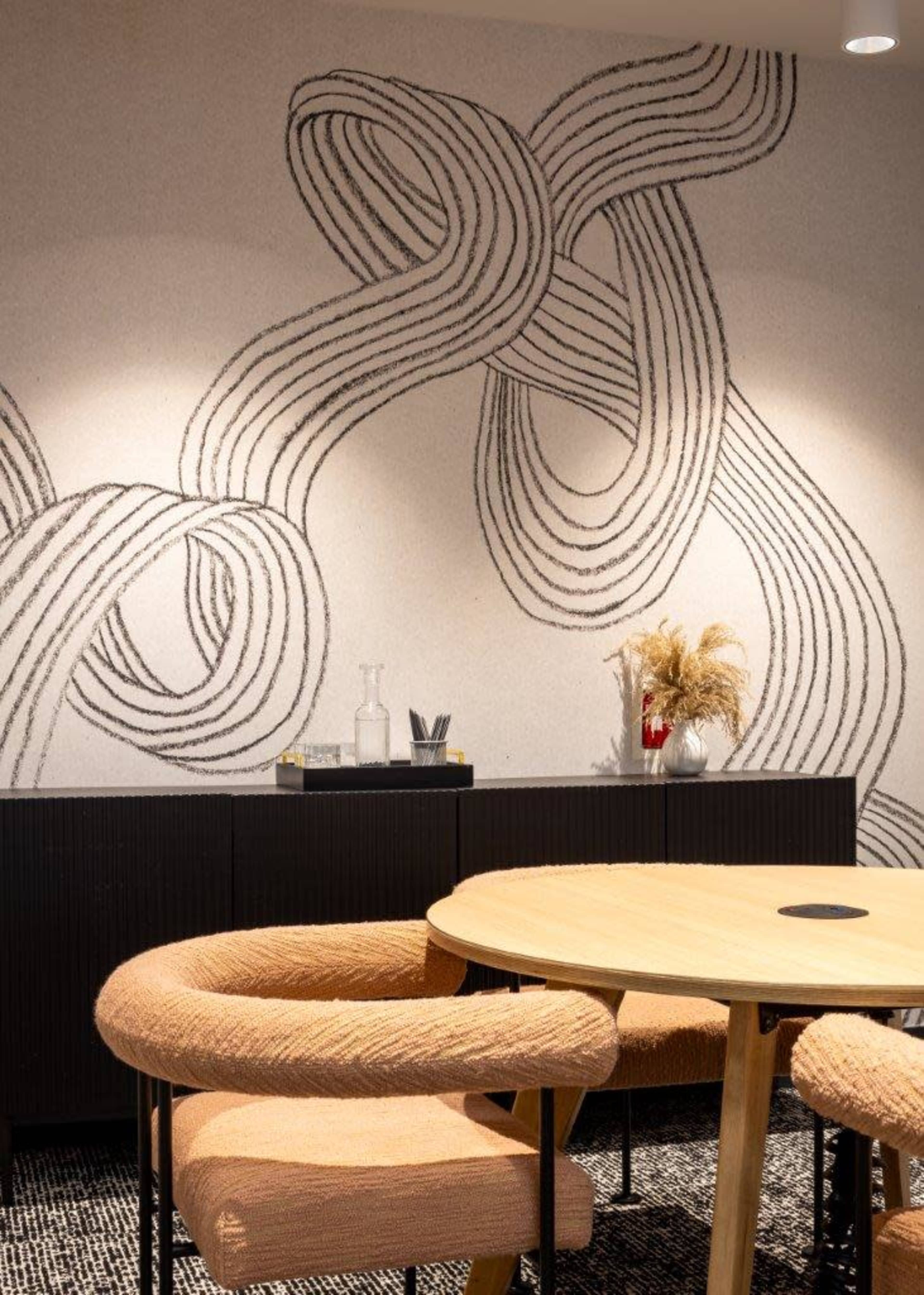 A modern interior featuring a round wooden table surrounded by upholstered chairs, with a decorative wall displaying abstract line art and a sideboard with decorative items.