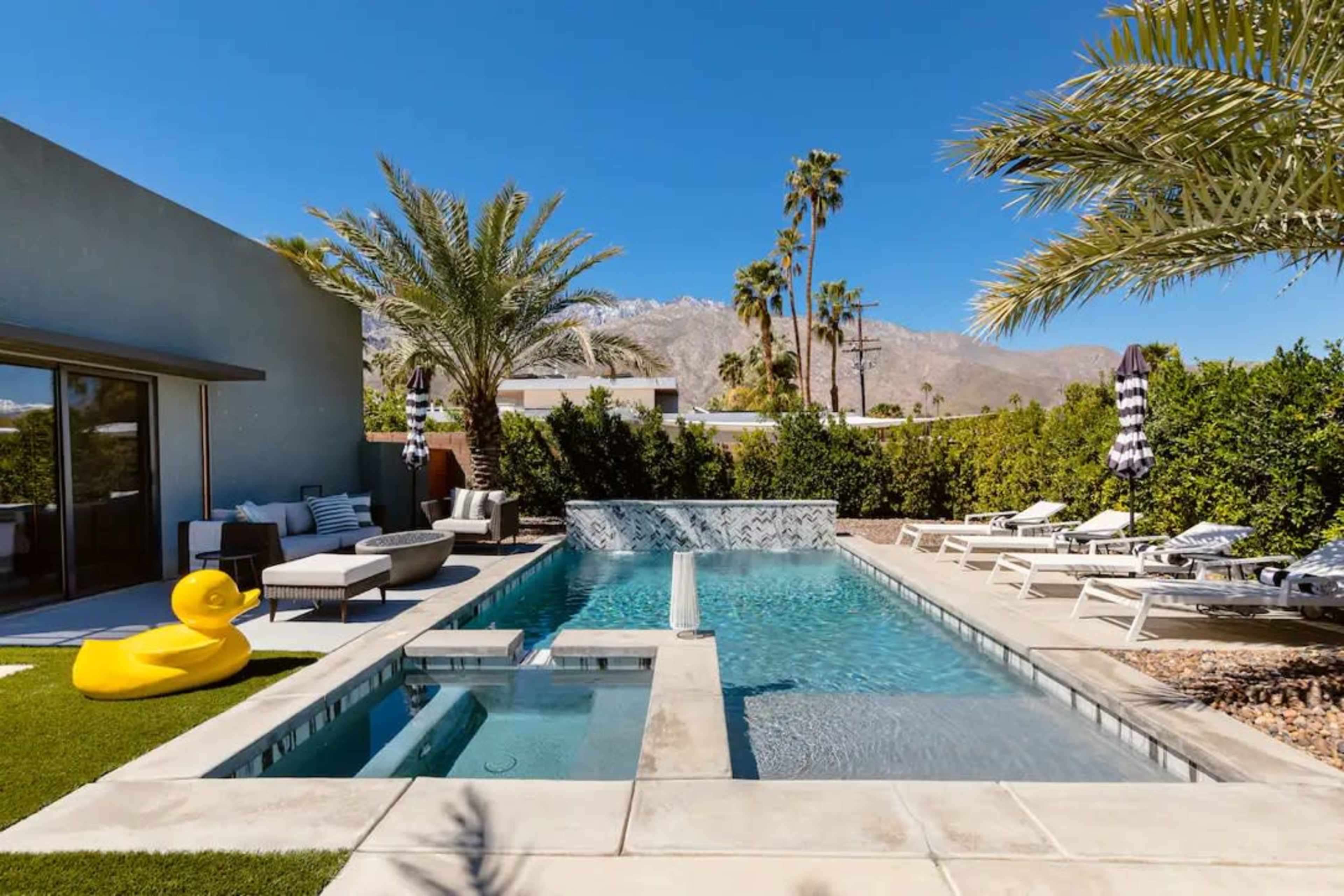 The image shows a modern backyard with a swimming pool, spa, palm trees, lounge chairs, and a large yellow rubber duck.