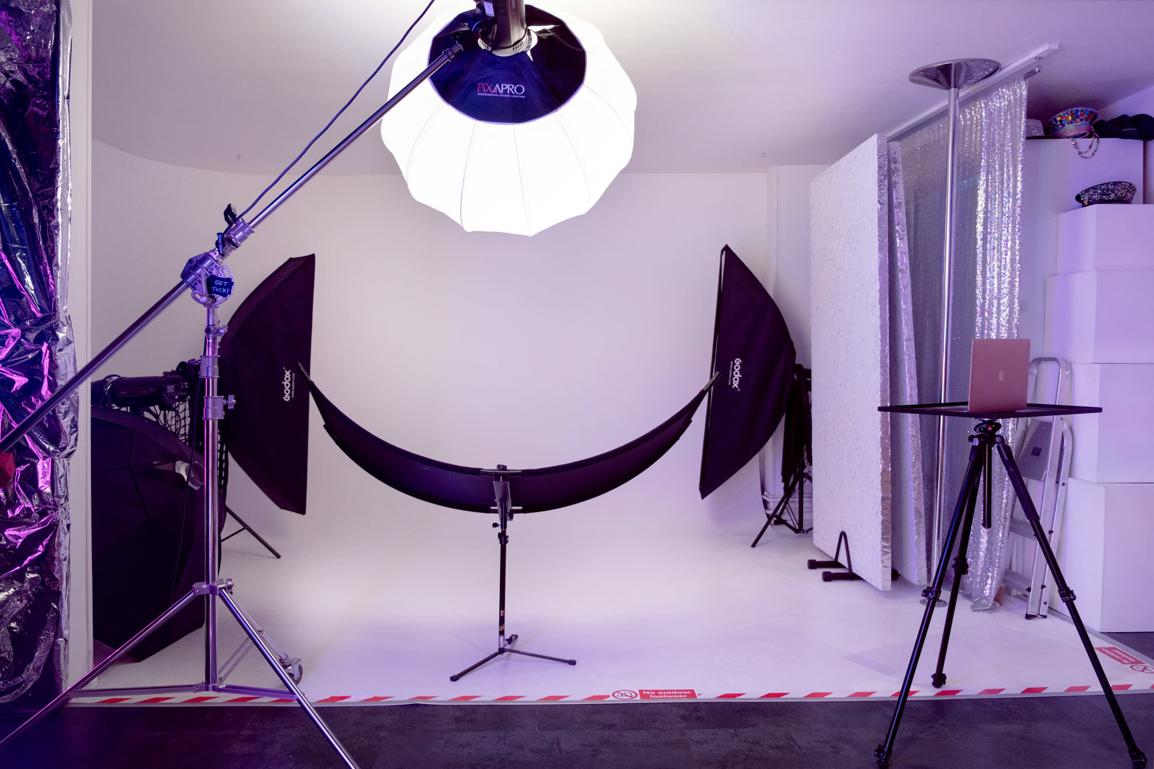 The image shows a photography studio setup with a white backdrop, two softbox lights, and a table with a laptop positioned nearby.
