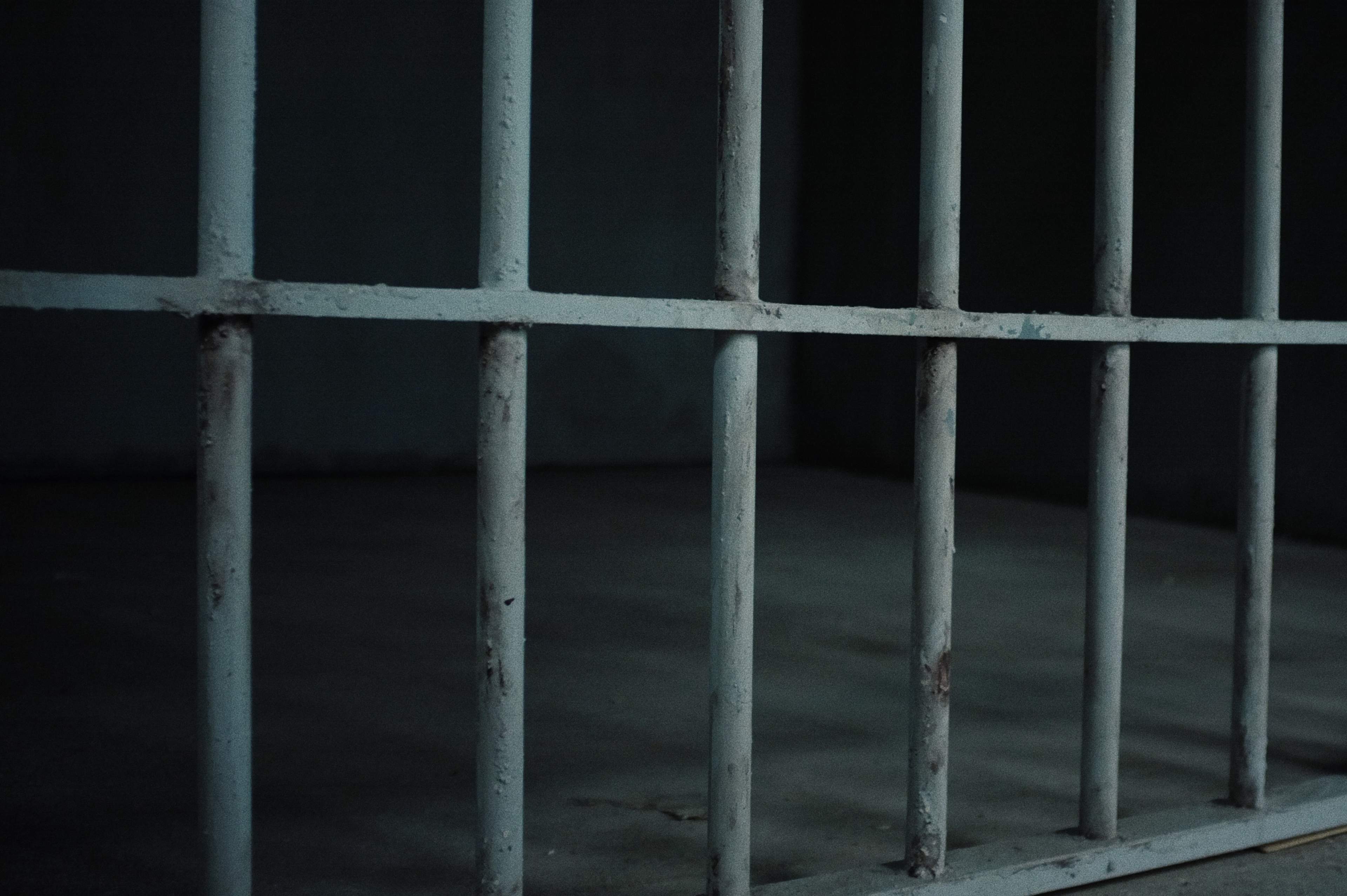 The image shows a close-up of rusted metal bars of a jail cell with a bare floor behind them.