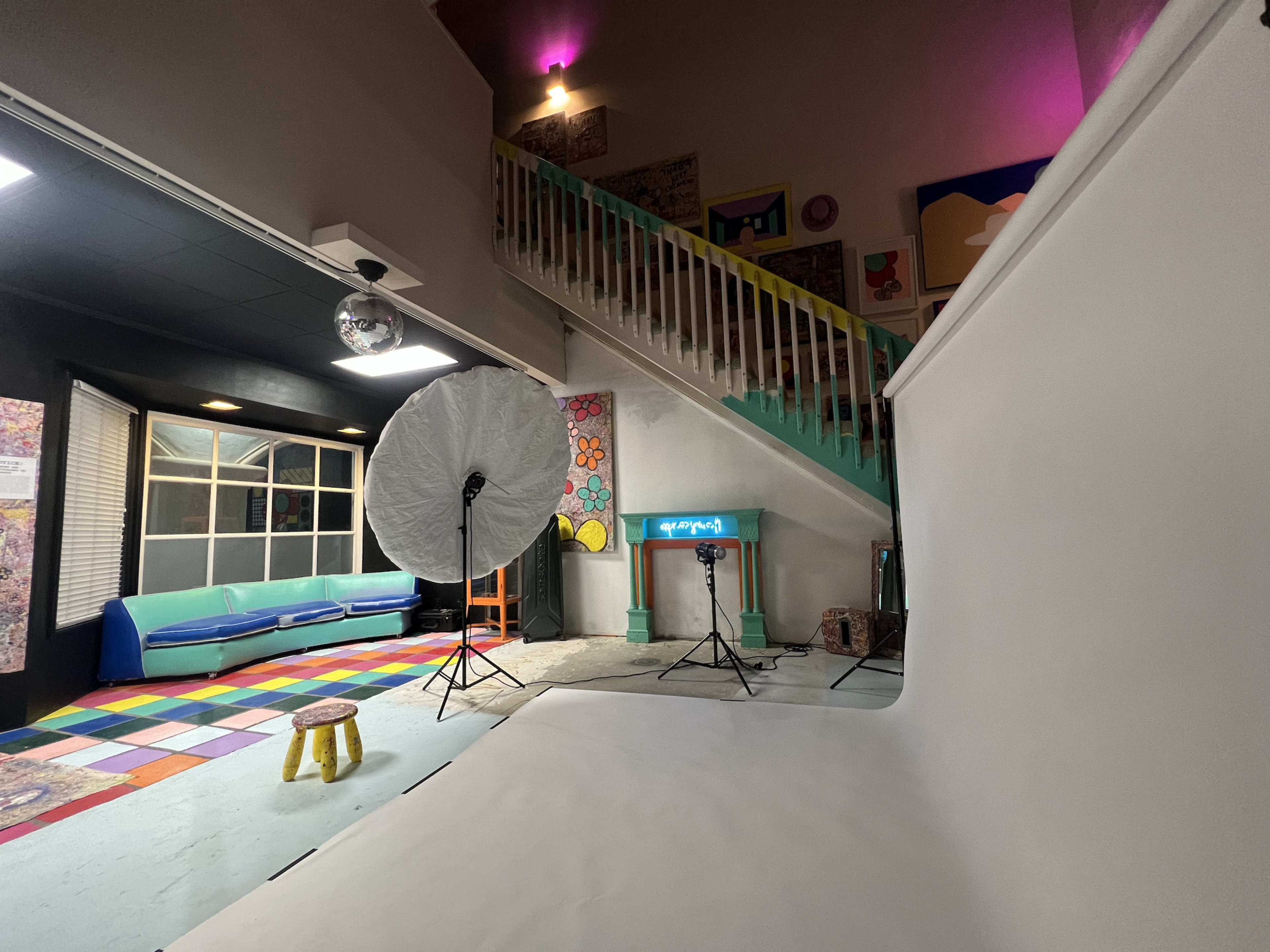 The image shows a vibrant, multi-colored studio space with a staircase, a large lighting setup, and eclectic artwork on the walls.