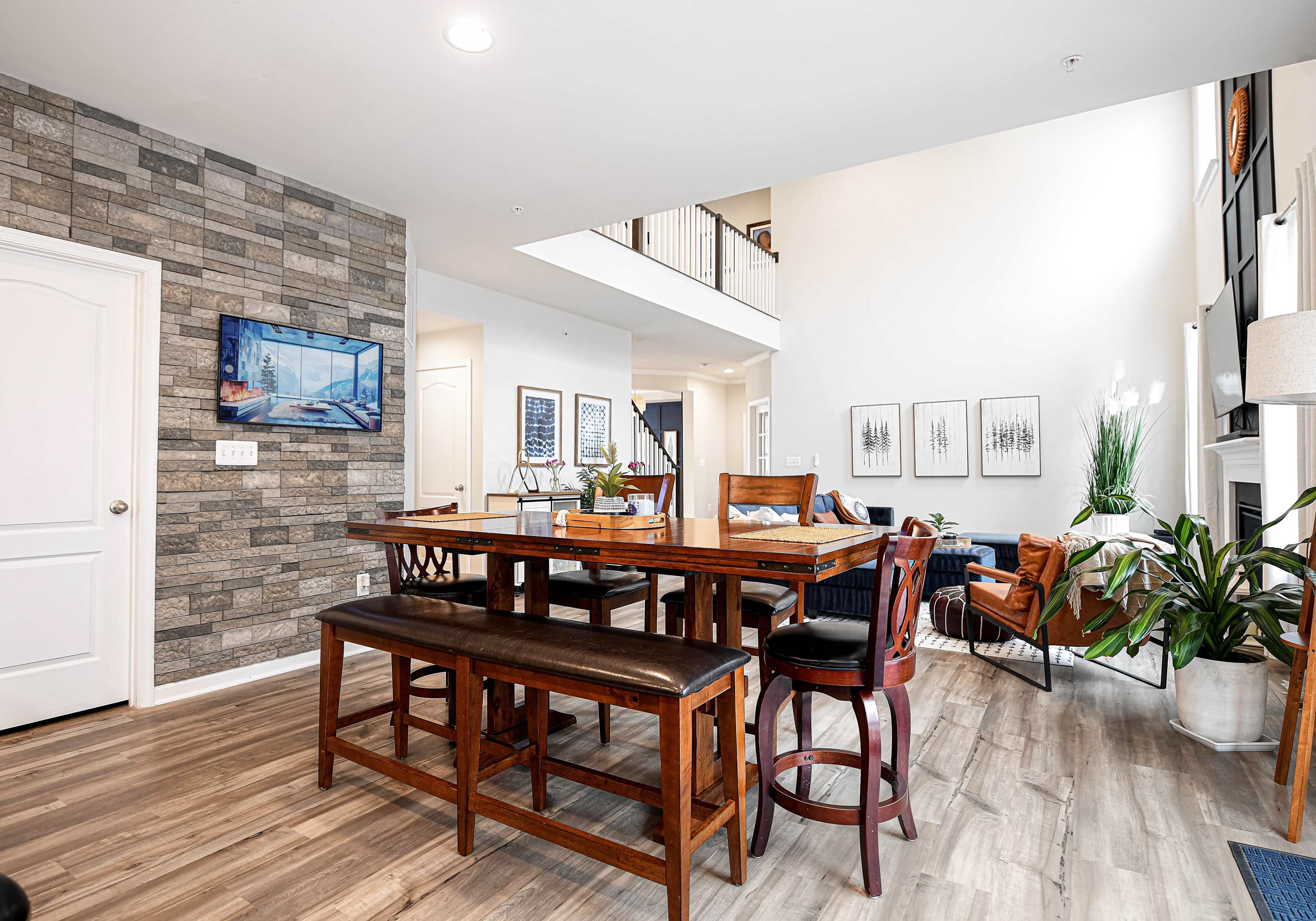 A spacious living area with a wooden dining table, seating, and a stone accent wall, featuring natural light from large windows.