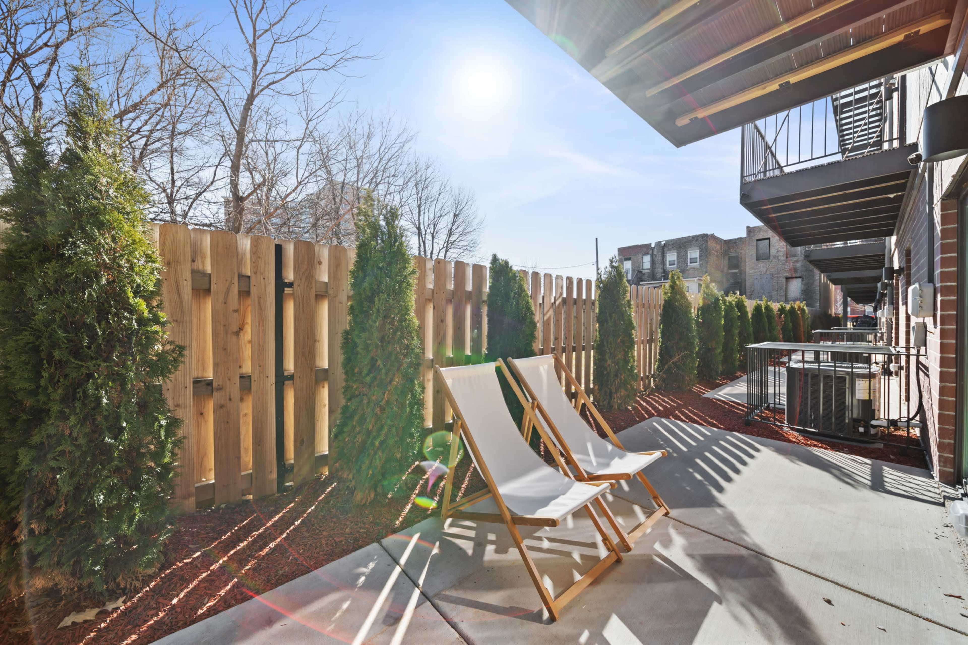 A private outdoor patio features two lounge chairs beside a wooden fence and lined with small trees.