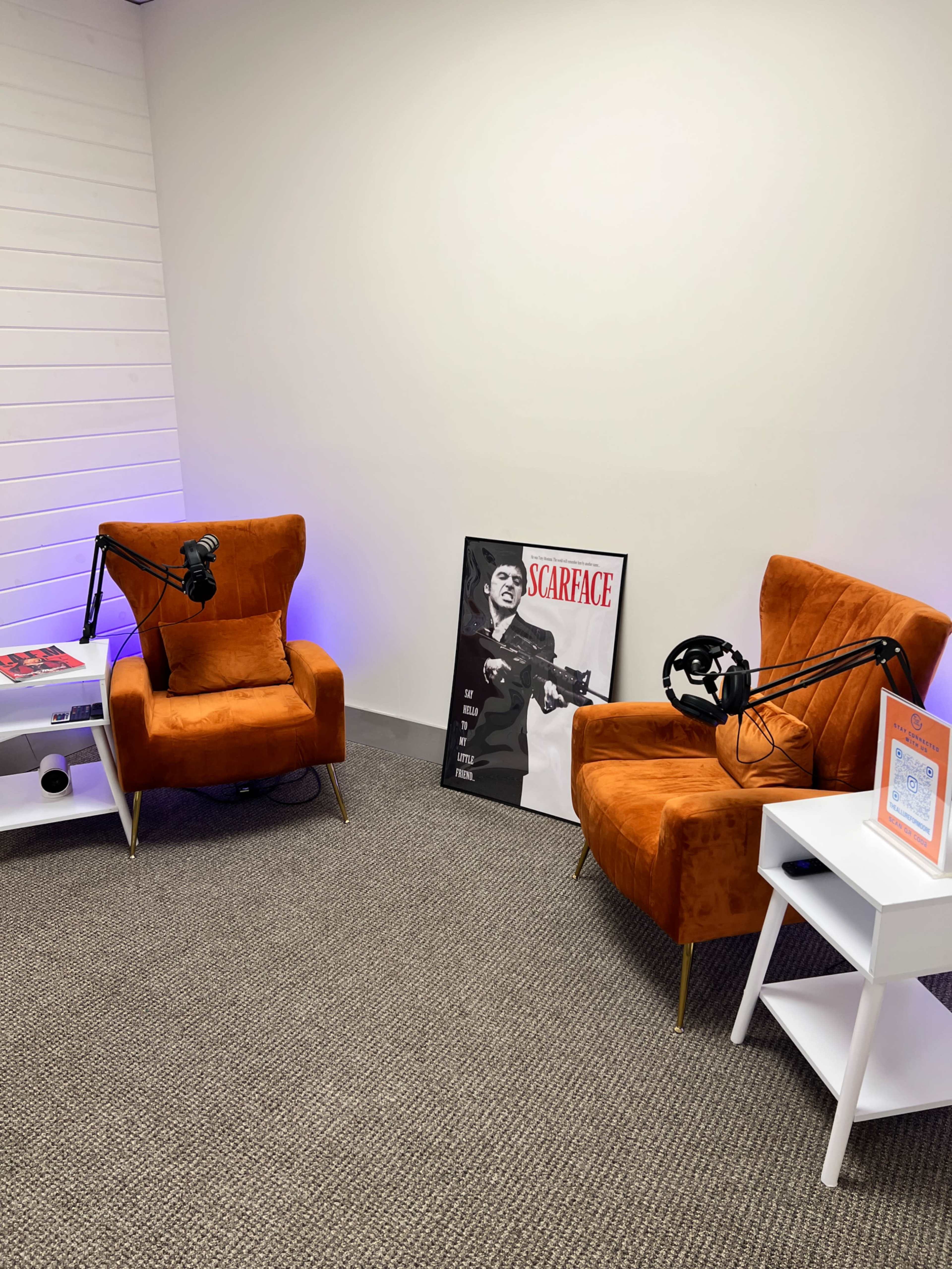 The image shows a small recording studio featuring two orange armchairs, a white side table, and a poster of "Scarface" on the floor, with microphones mounted above the chairs.