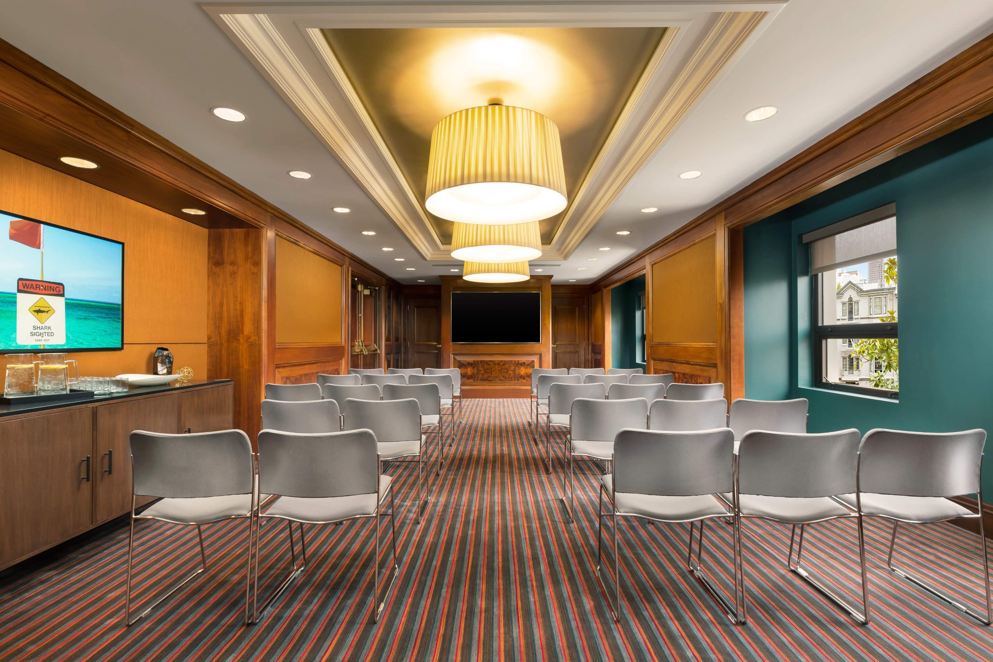 The image shows a conference room with rows of empty chairs facing a large screen and large light fixtures overhead.