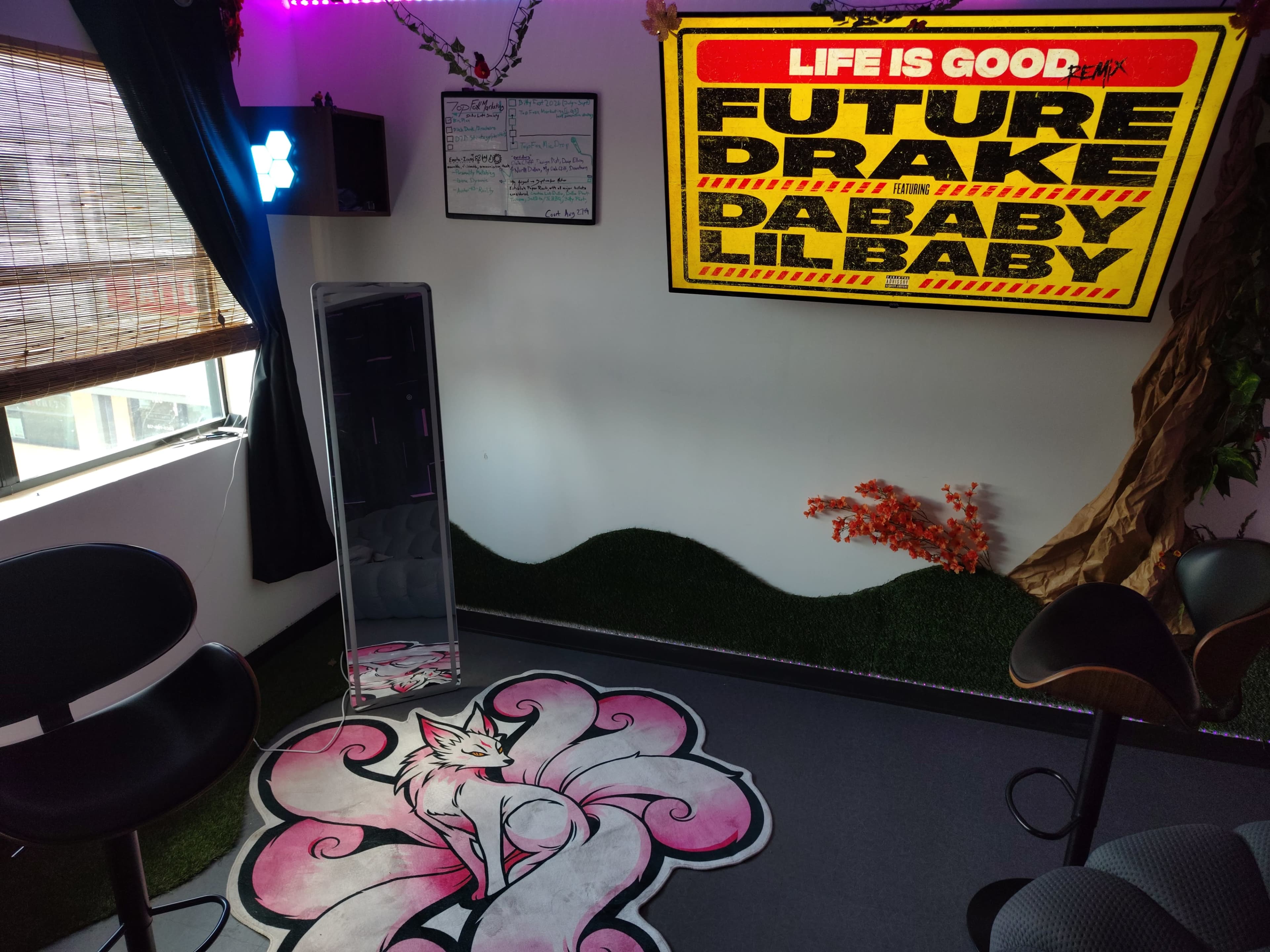 The image shows a corner of a room with a large wall-mounted screen displaying music-related graphics, a pink and white fox design on the floor, and modern black chairs.