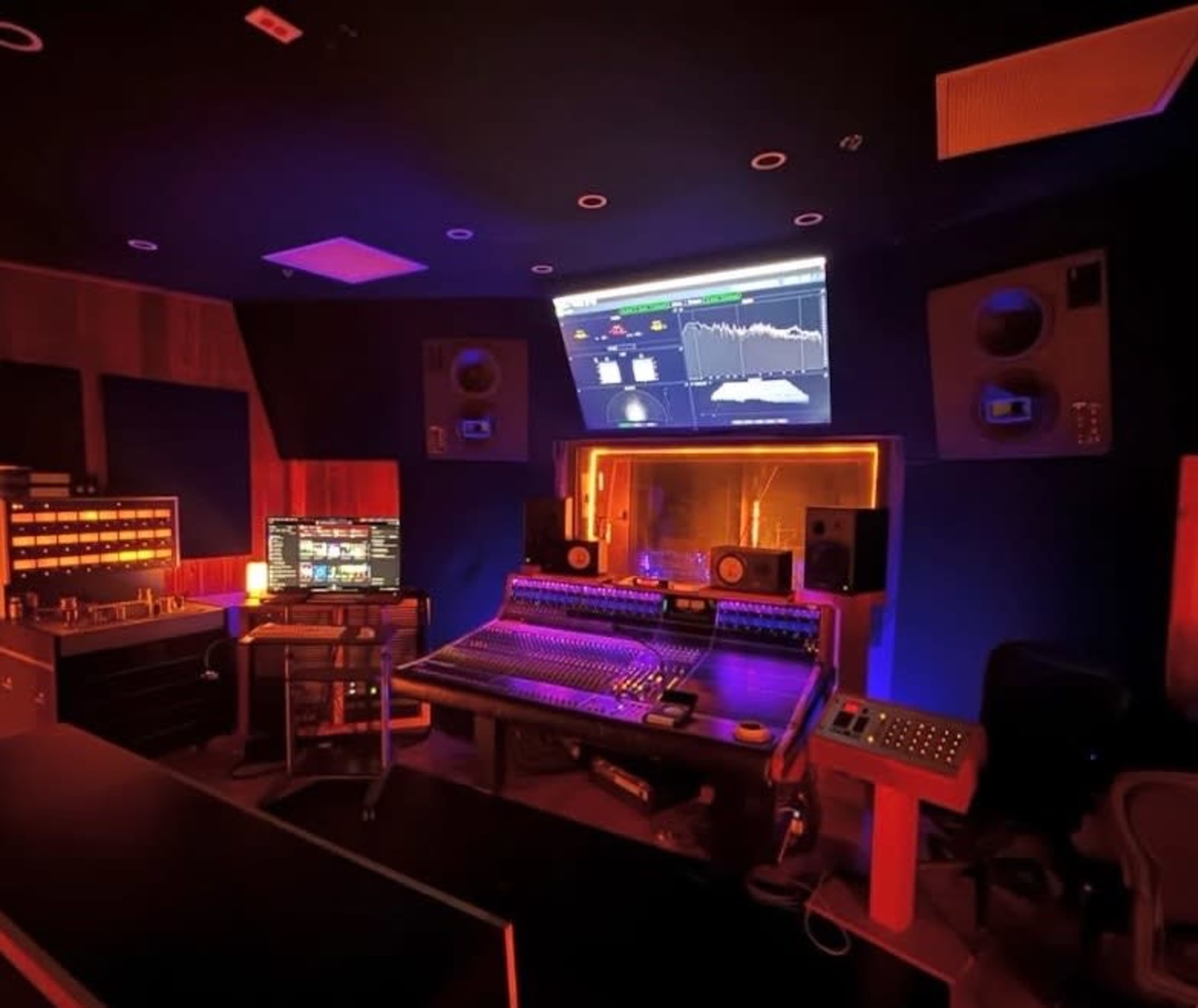 The image depicts a modern audio recording studio with a large mixing console, sound monitors, and a wall-mounted screen displaying audio levels and graphs.