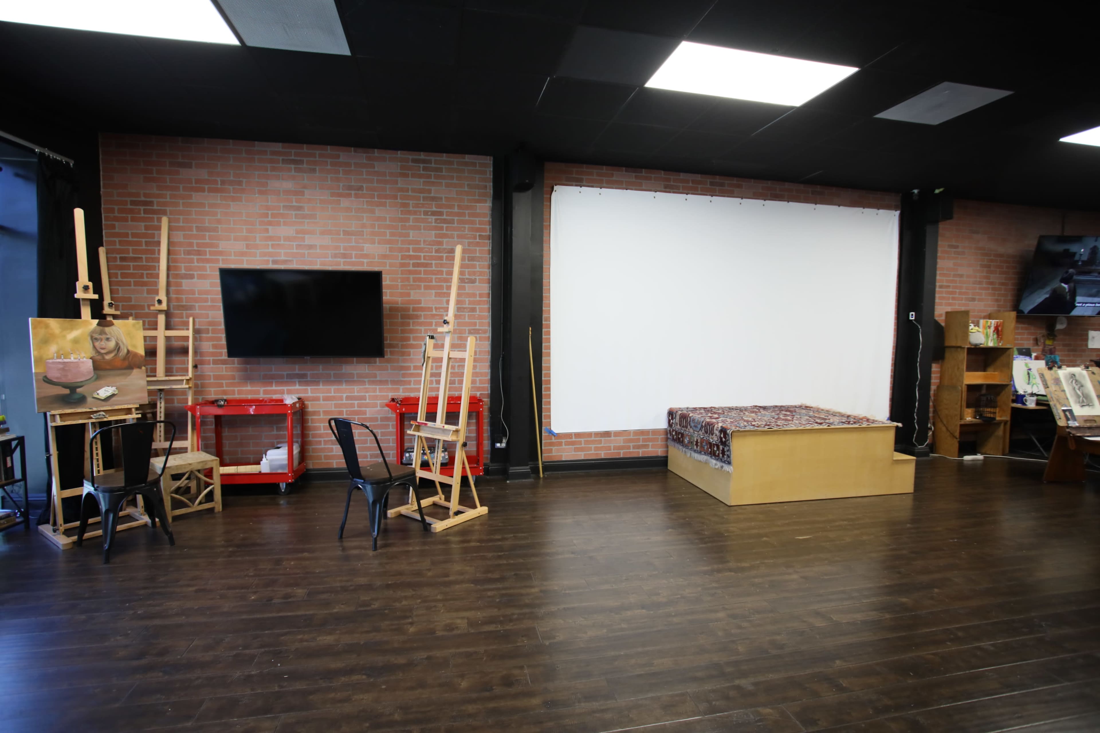 The image shows an art studio featuring easels, a seating area, a large blank screen, and a wooden platform with a decorative cover against a brick wall.
