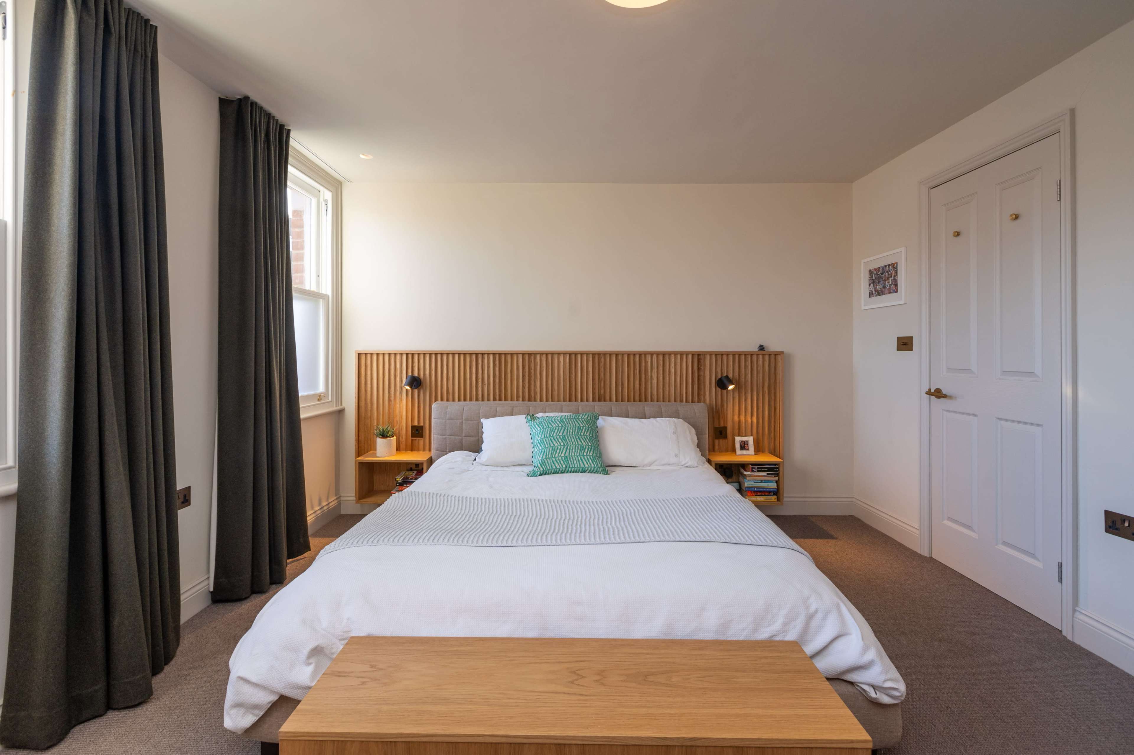 The image shows a tidy bedroom featuring a double bed with a wooden headboard, two bedside lamps, curtains, and a small wooden table at the foot of the bed.