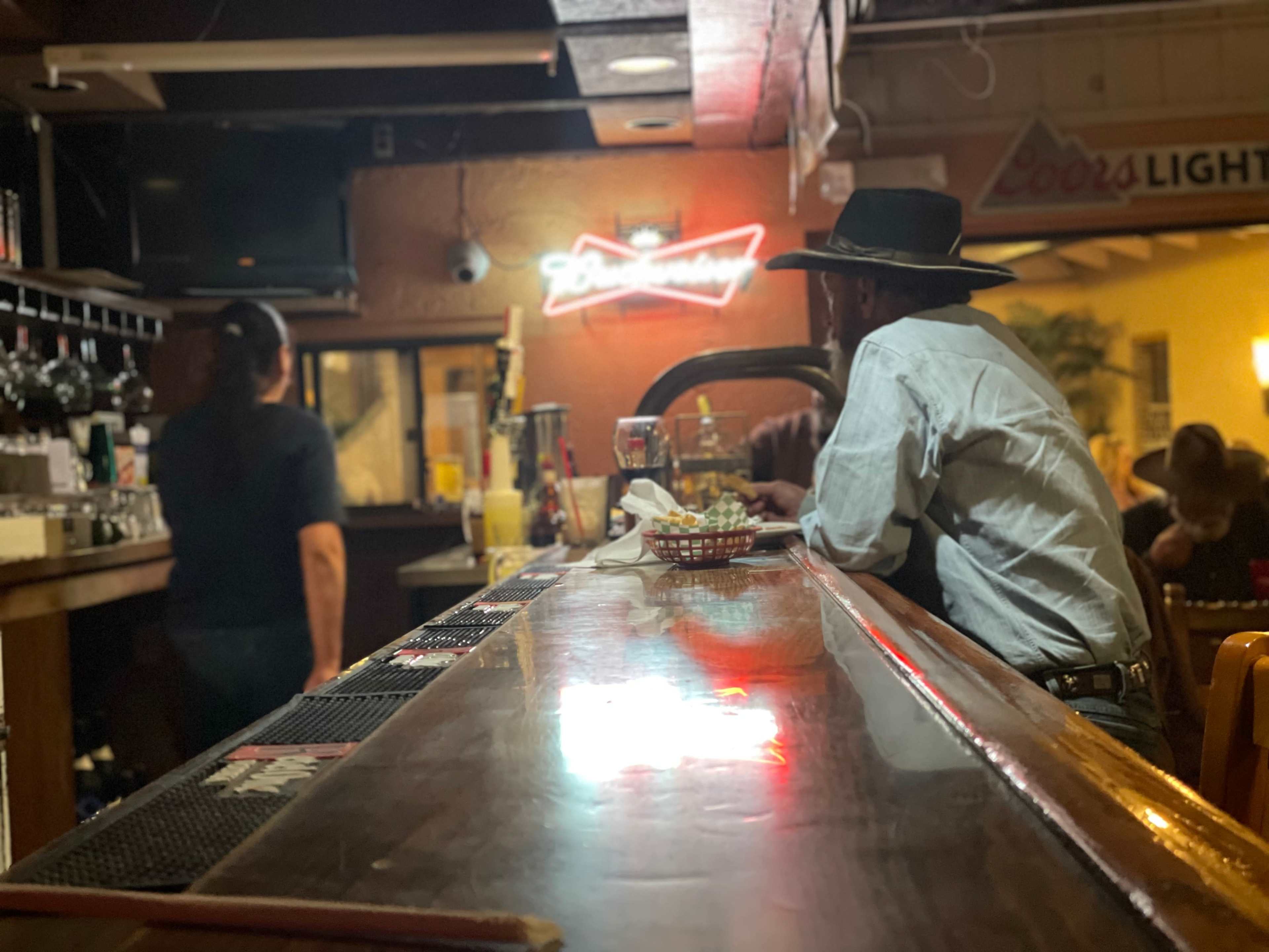 A man wearing a cowboy hat sits at the bar while a bartender prepares drinks in a dimly lit bar.