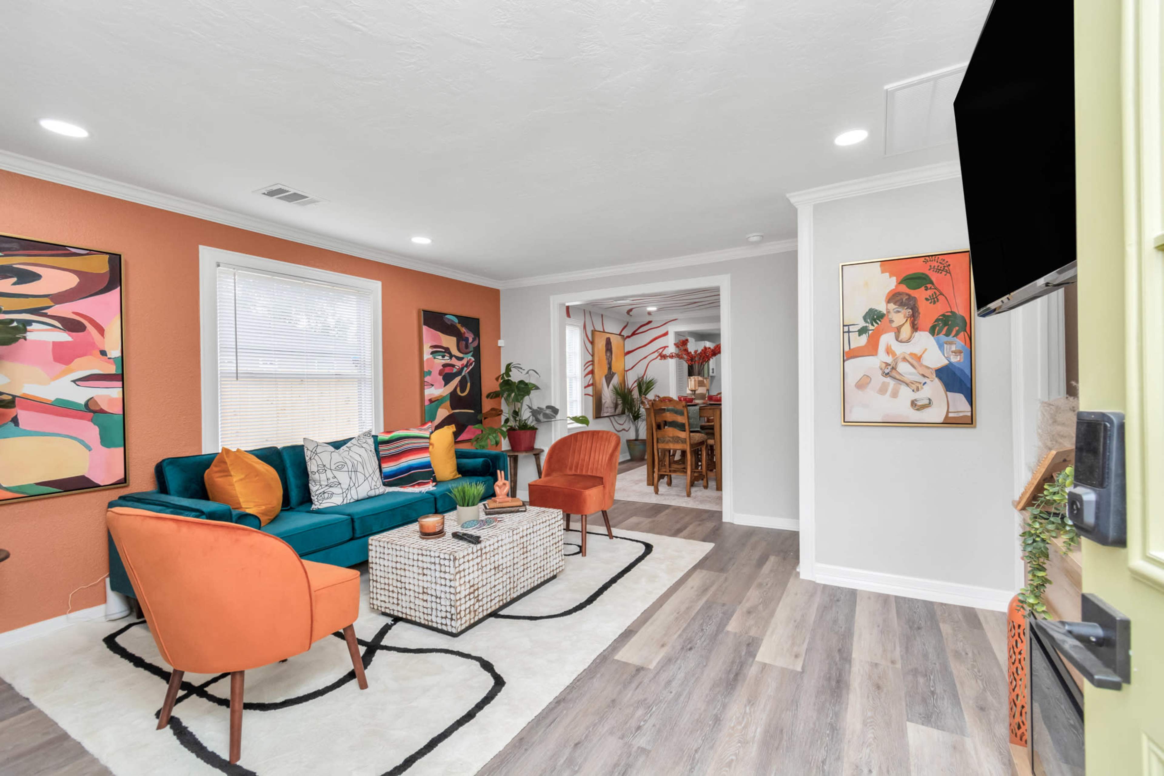 A bright living room featuring a teal sofa, orange accent chairs, a patterned coffee table, and colorful wall art, with a view into a dining area through an archway.