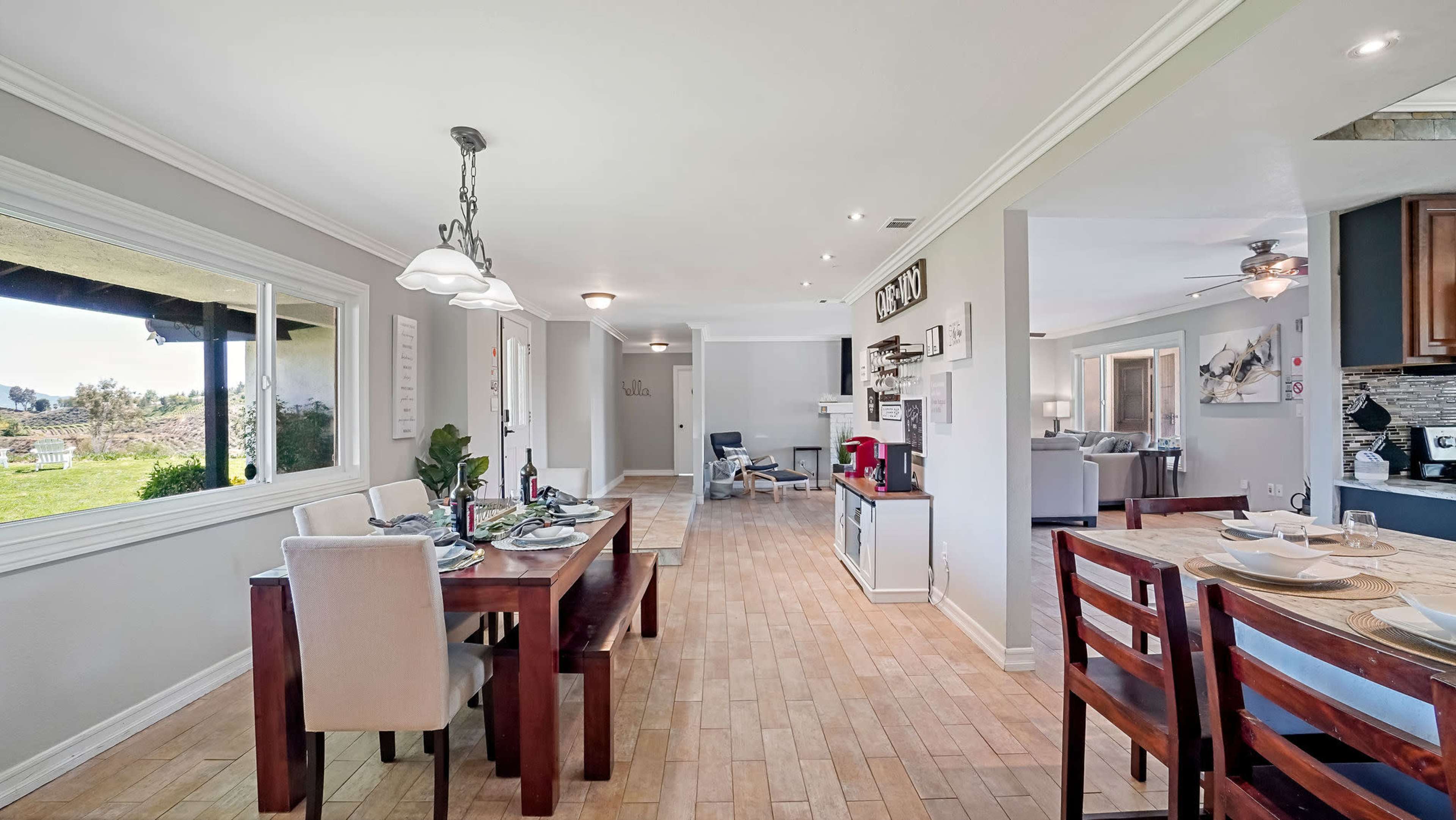 A dining area with a wooden table set for a meal, adjacent to a living space with a couch and TV, illuminated by natural light from large windows.