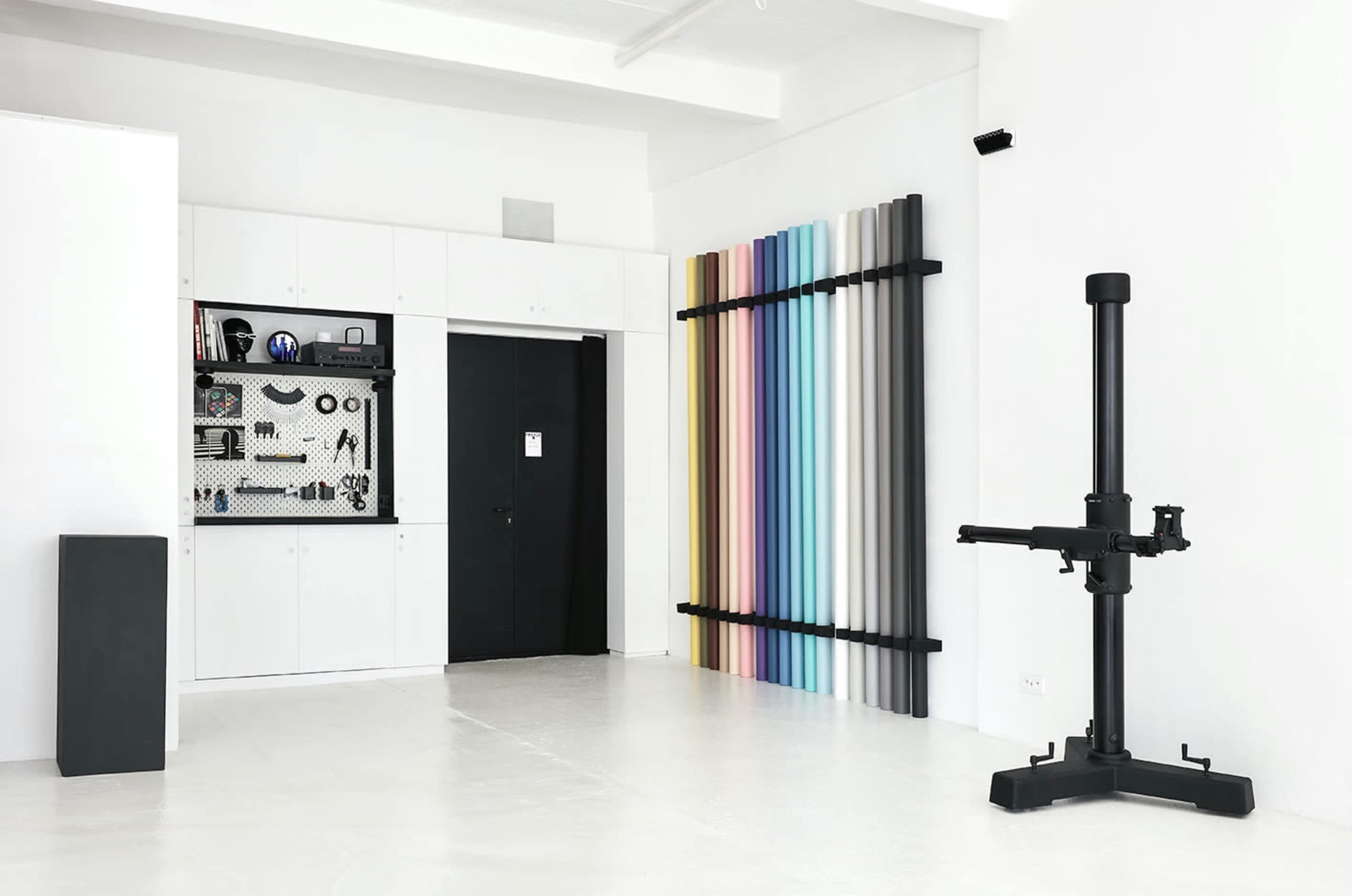 A modern studio space with a wall-mounted pegboard displaying tools, a door, a color panel with vertical stripes, and a large black filmmaking equipment stand.