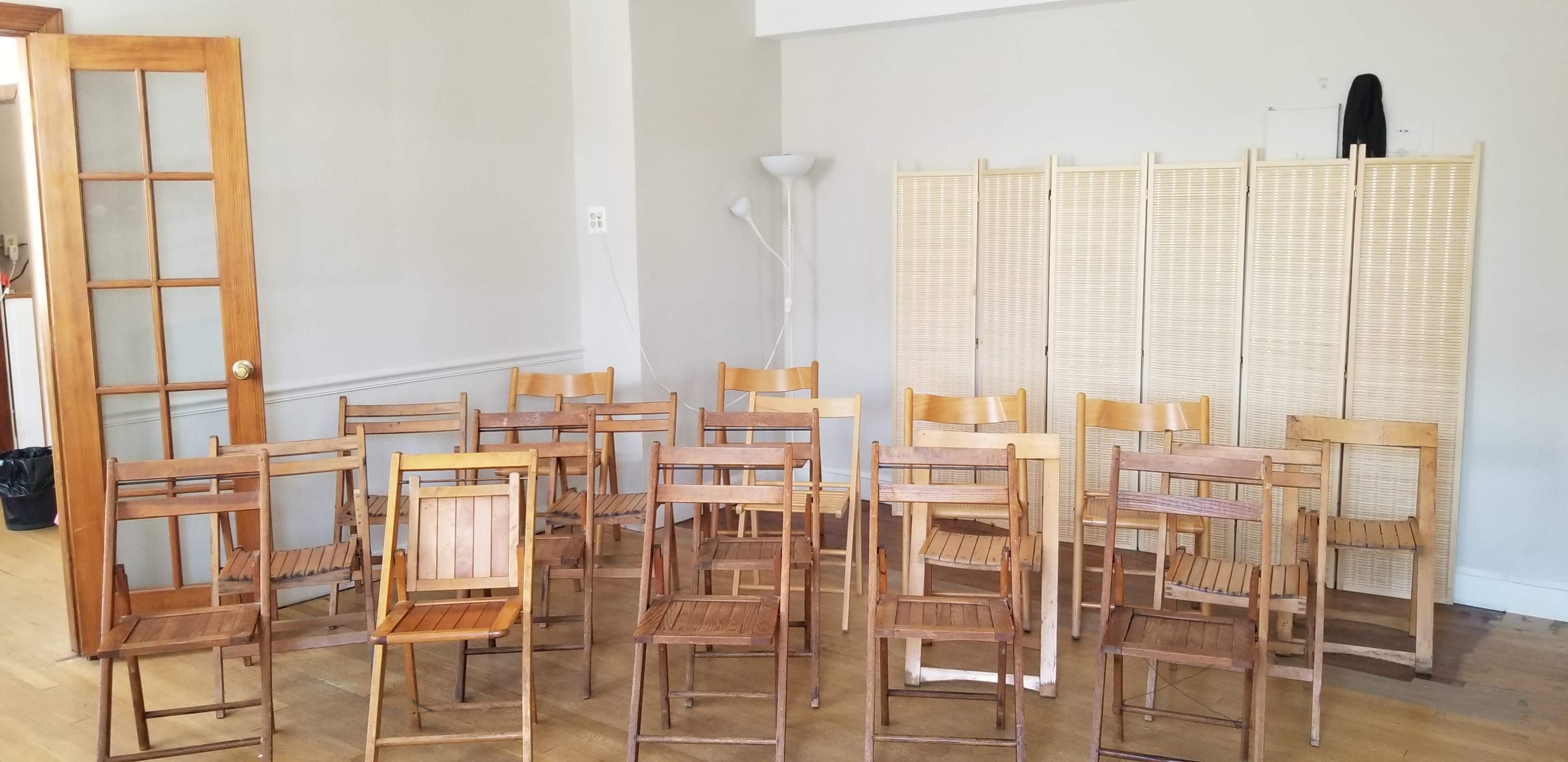 A group of wooden folding chairs is arranged in a room beside a folding screen and a lamp.