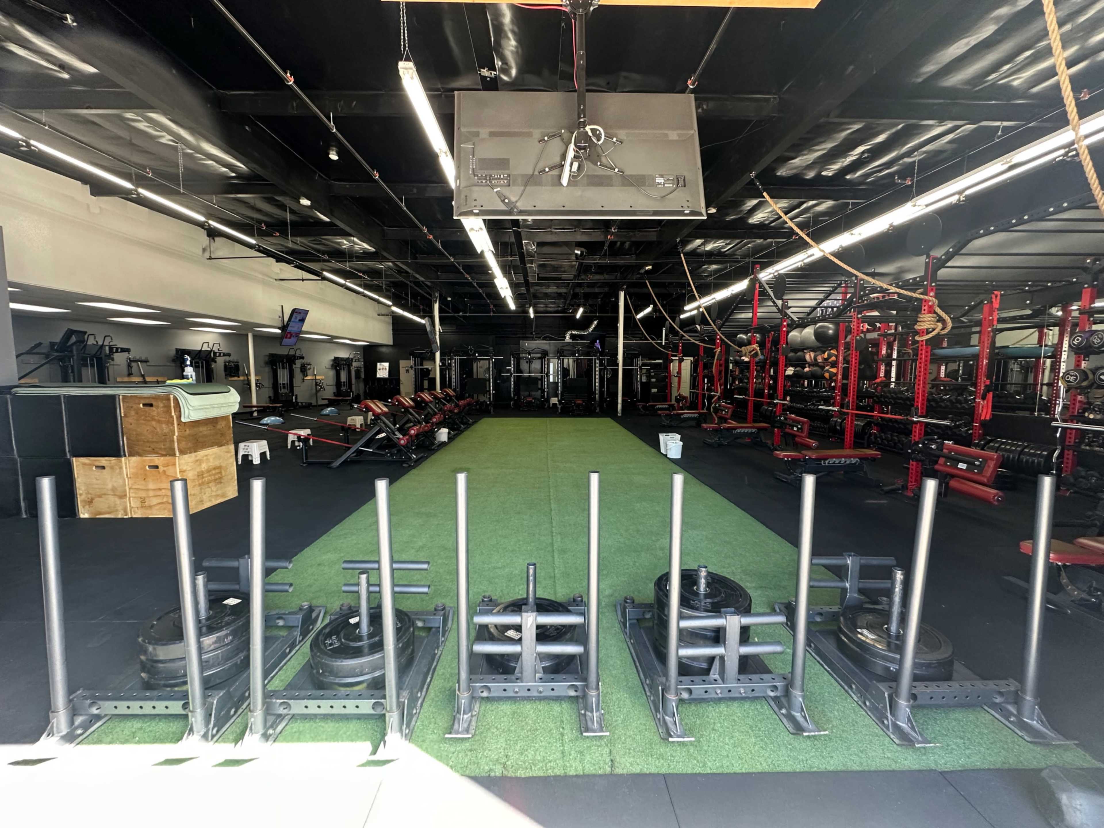 The image shows a spacious gym filled with various weightlifting equipment, including sleds at the front and rows of benches and machines in the background.