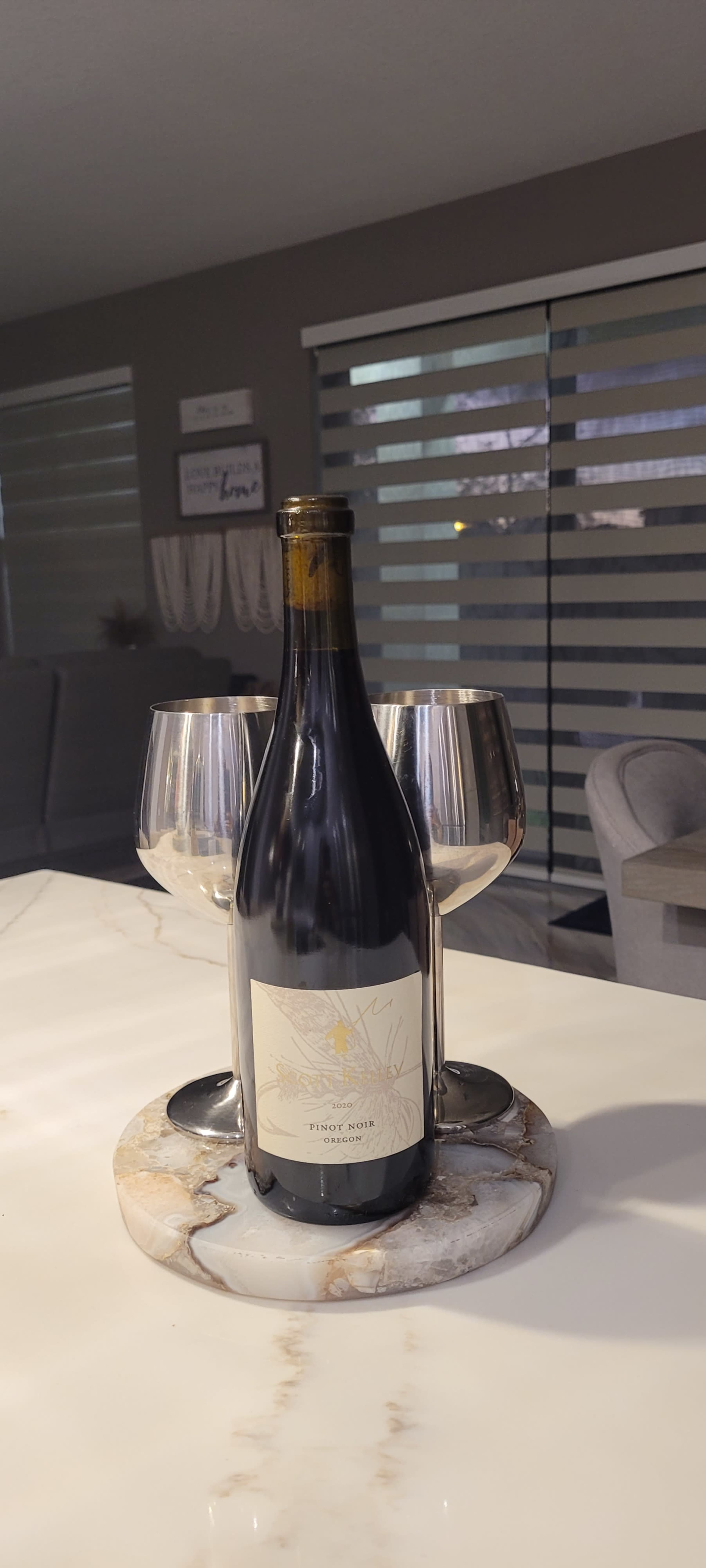 A bottle of Pinot Noir is placed on a circular marble coaster between two wine glasses on a table.