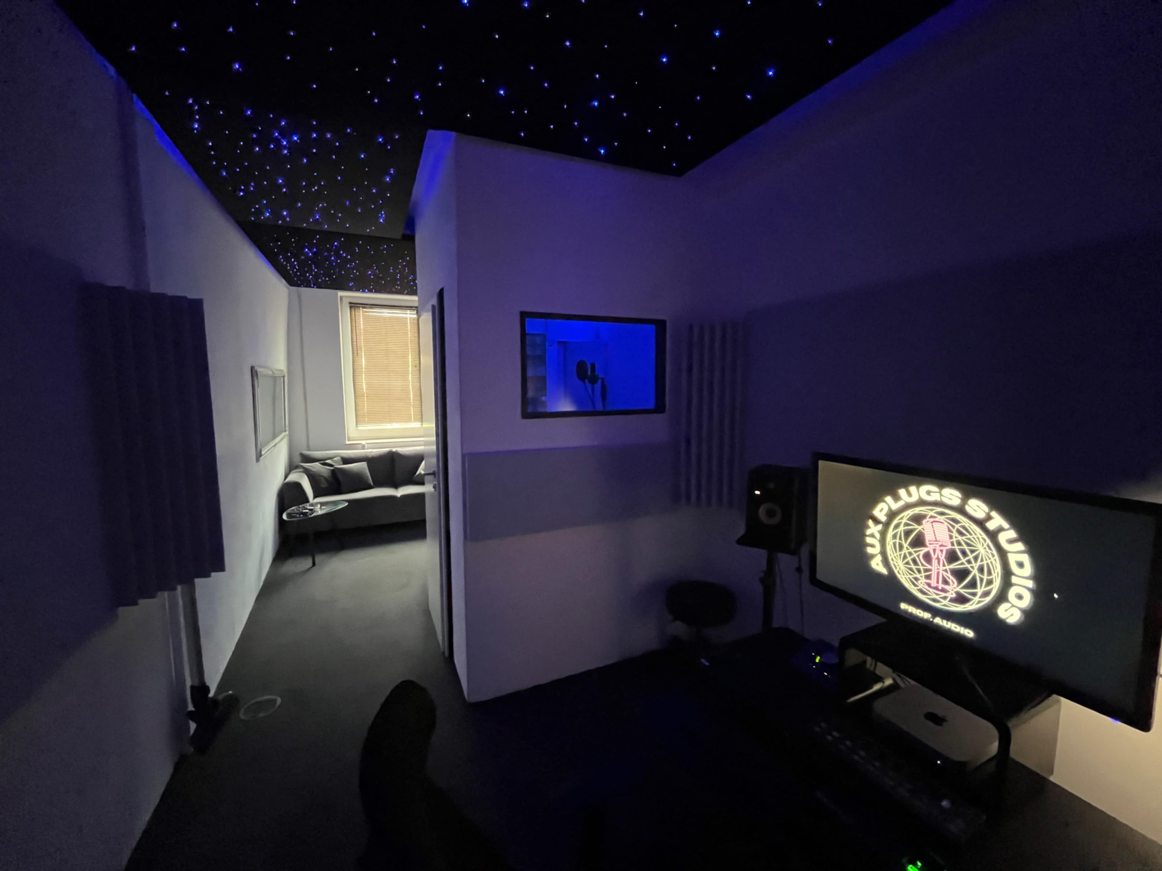 A dimly lit recording studio with a starry ceiling, a microphone in a soundproof booth, and a workspace featuring a computer and studio monitors.