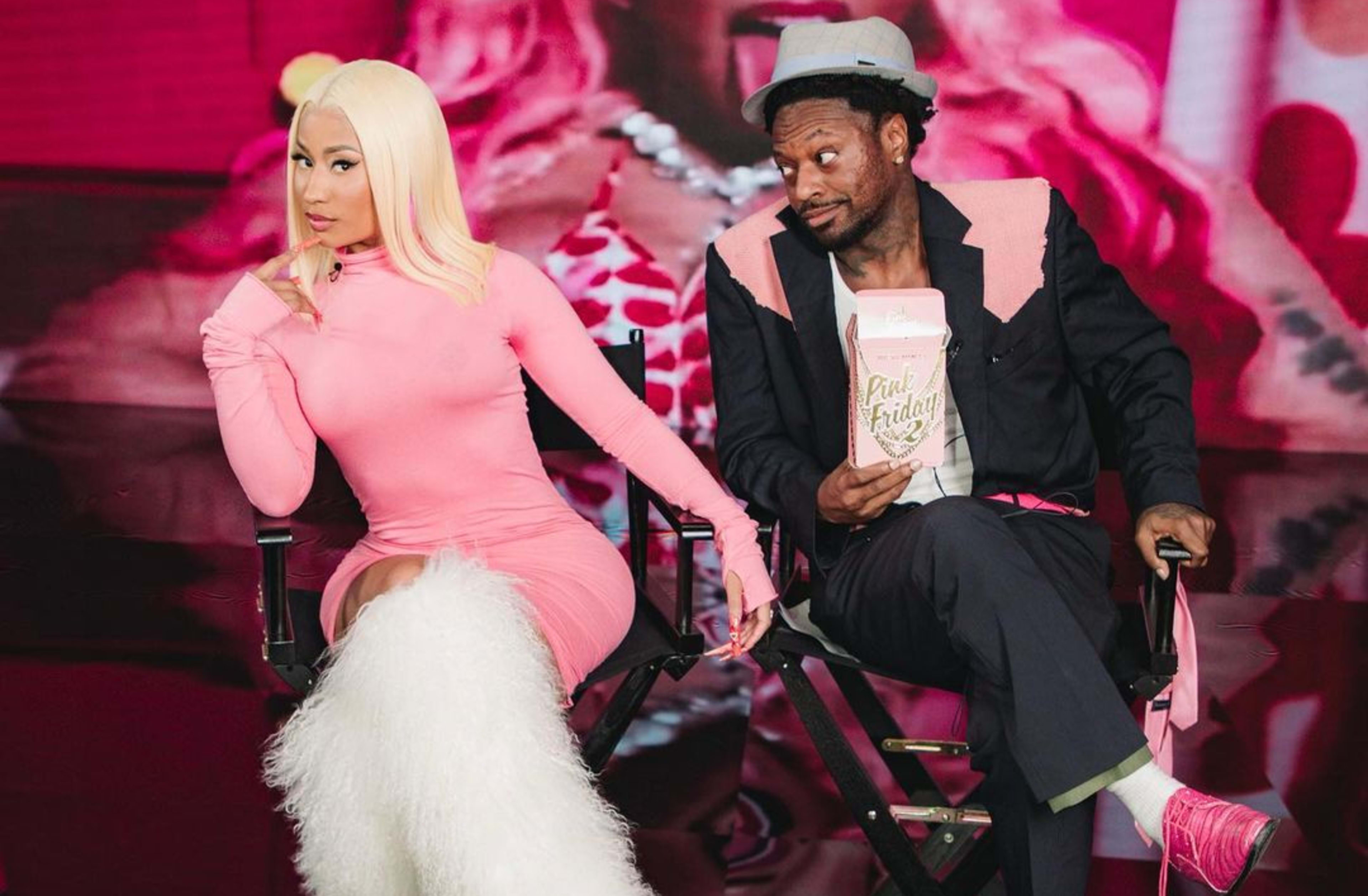 Nicki Minaj and a man are seated on director's chairs in a colorful, stylized setting, with the man holding a box labeled "Pink Friday."