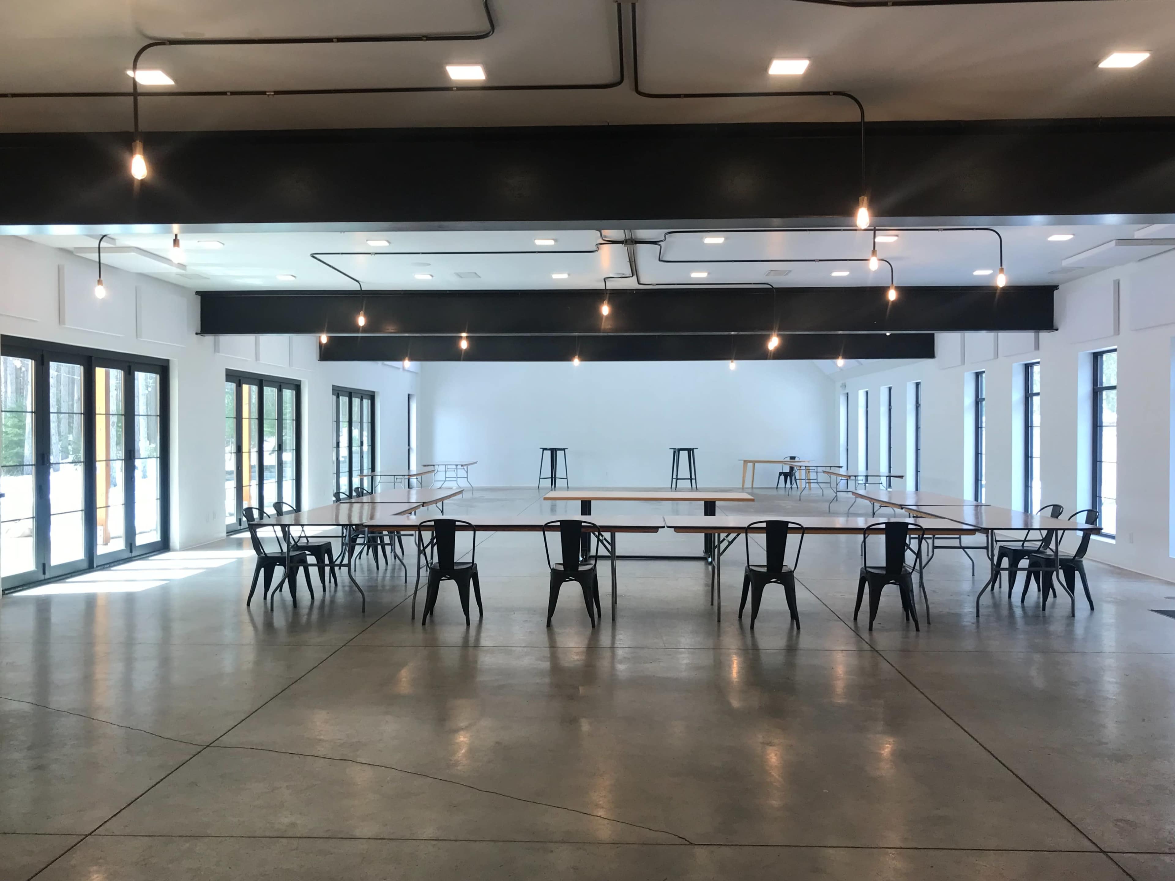 An empty, modern room with several tables arranged in a grid, surrounded by black chairs, and well-lit by hanging lights.