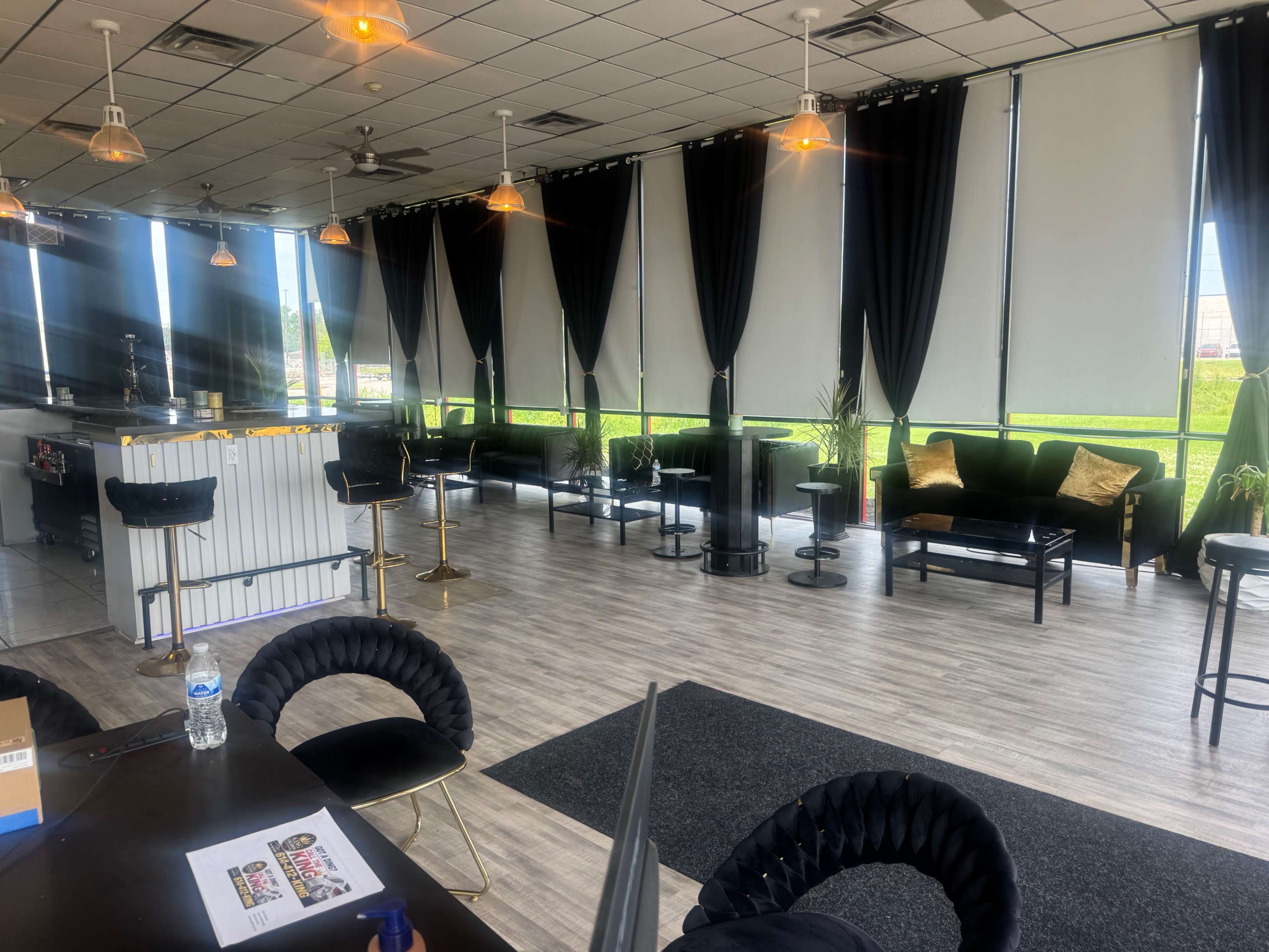 The image shows a modern lounge area with black curtains, black furniture, and large windows allowing natural light to enter.