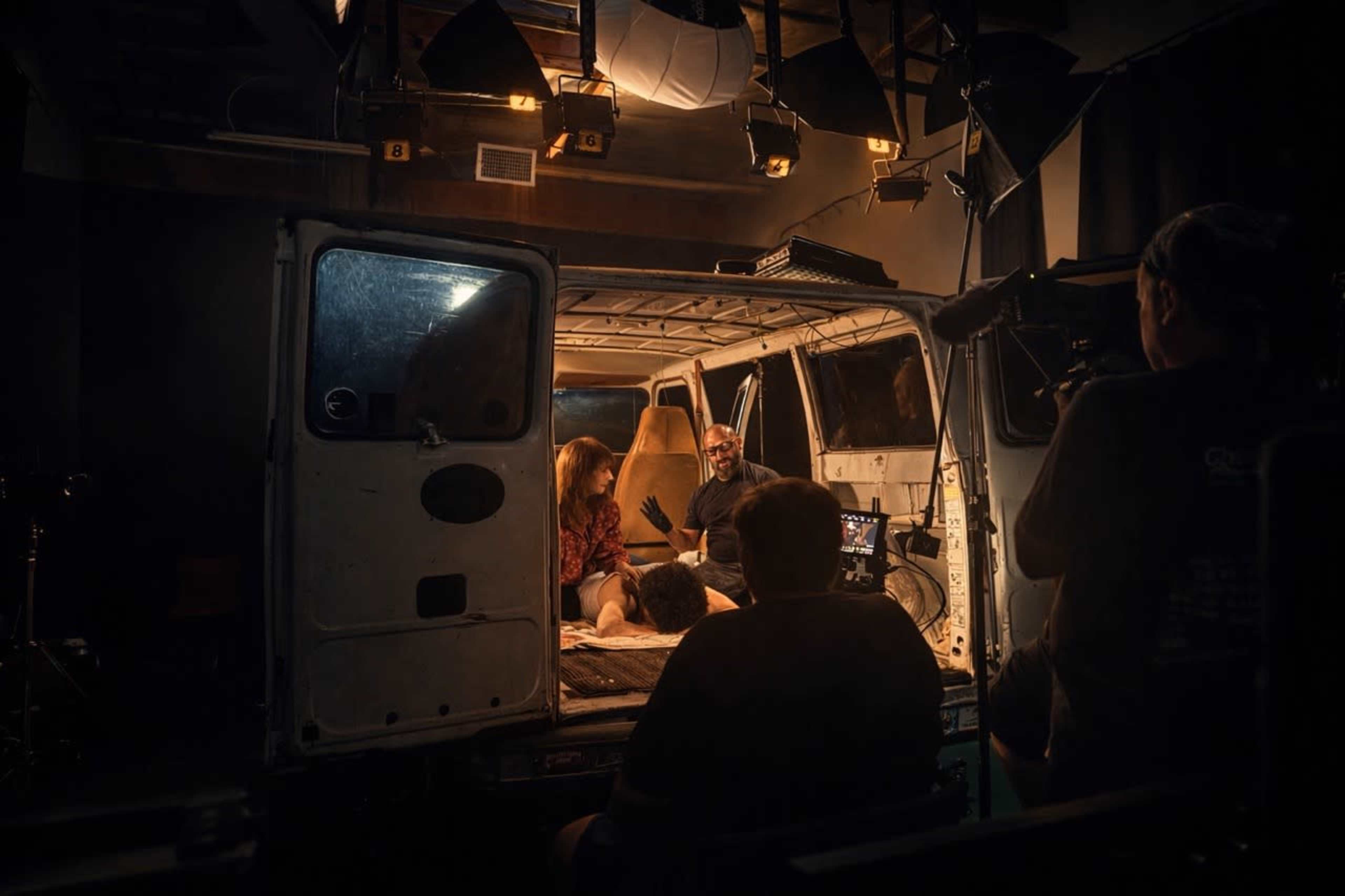 A film crew is inside a van setting up a scene with two actors engaged in conversation under bright studio lights.