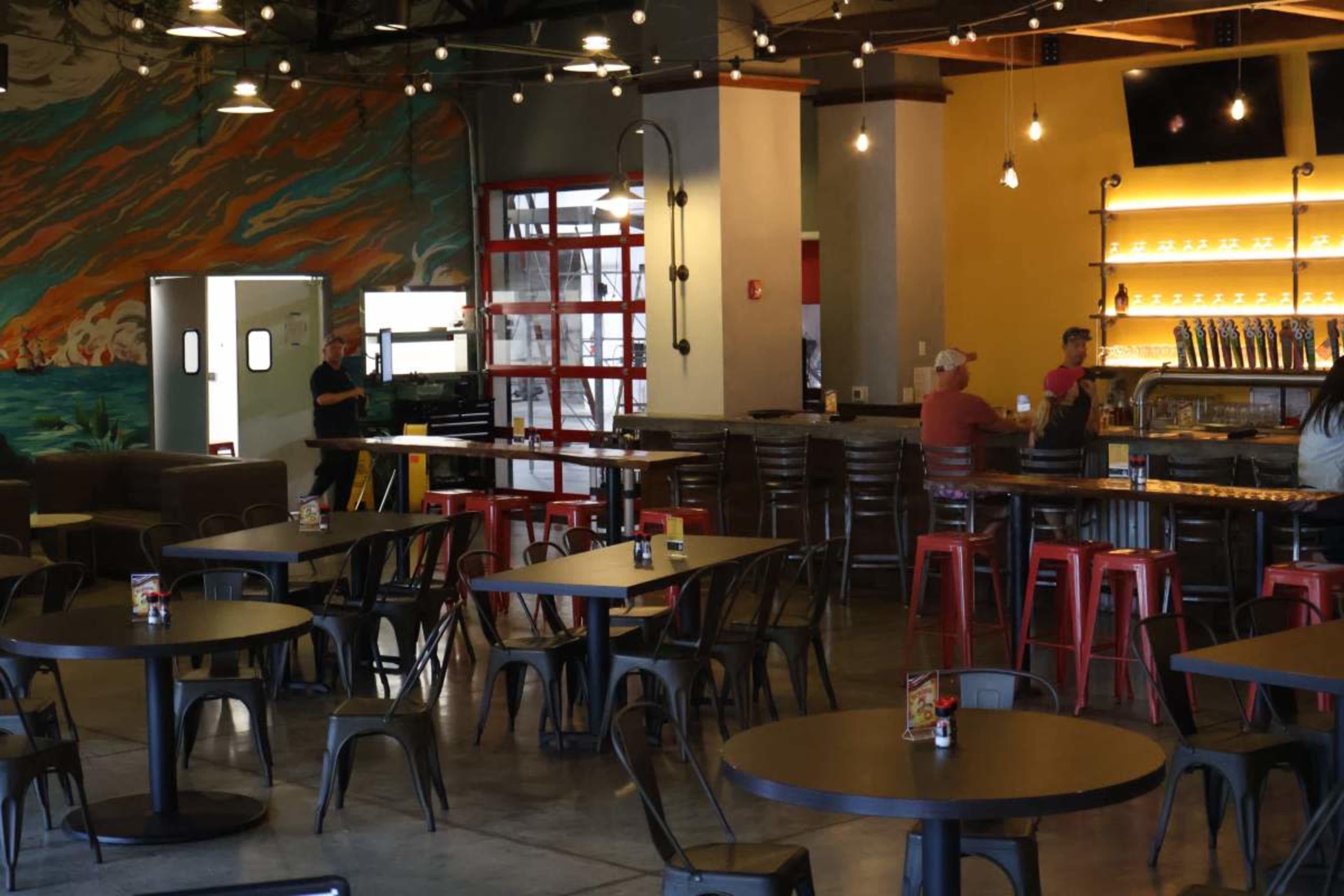 The scene depicts a modern restaurant interior with metal tables and chairs, a bar area with illuminated shelves, and a vibrant mural on the wall.