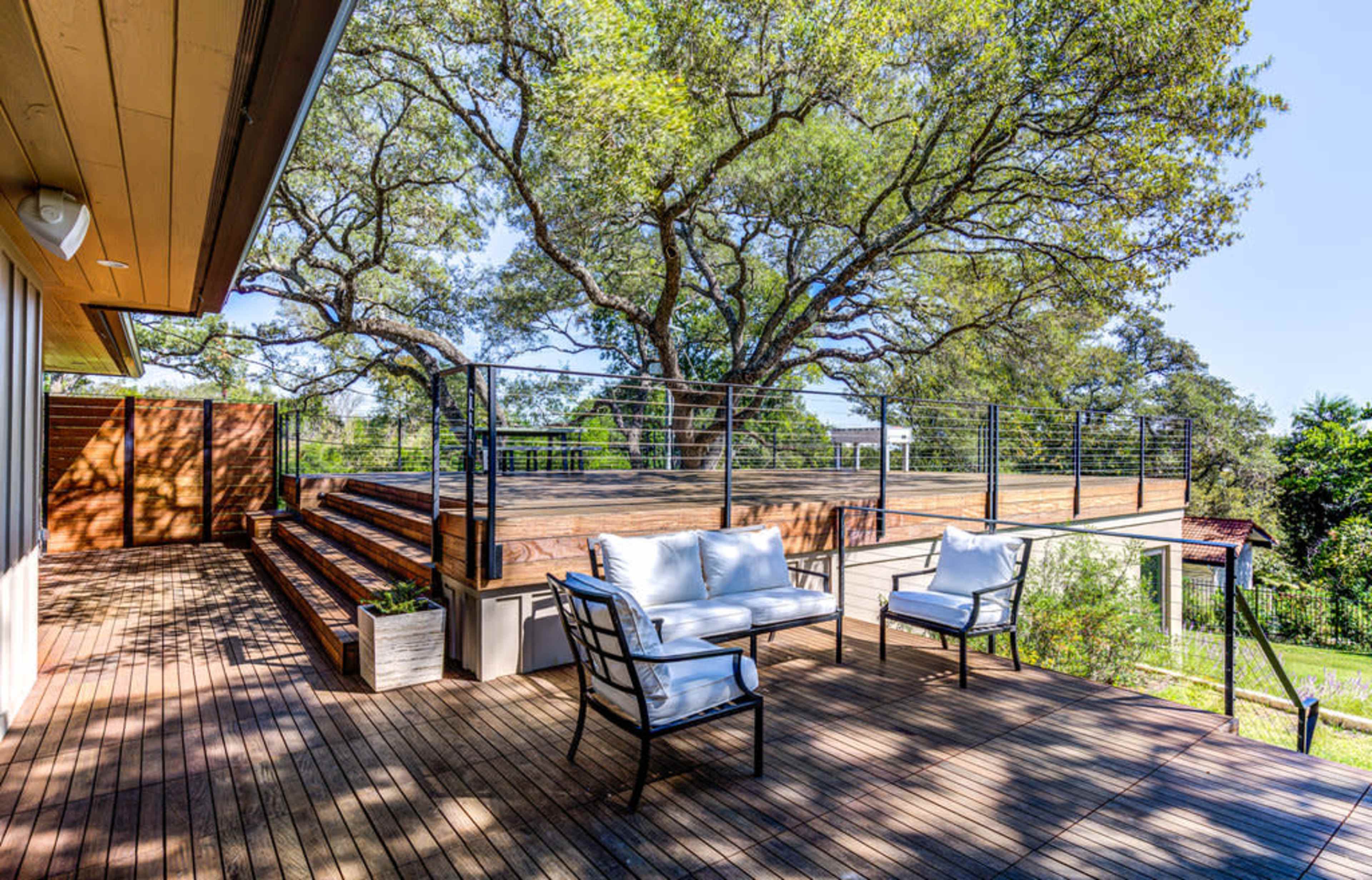 A wooden deck with seating overlooks a landscaped area and large trees under bright blue skies.