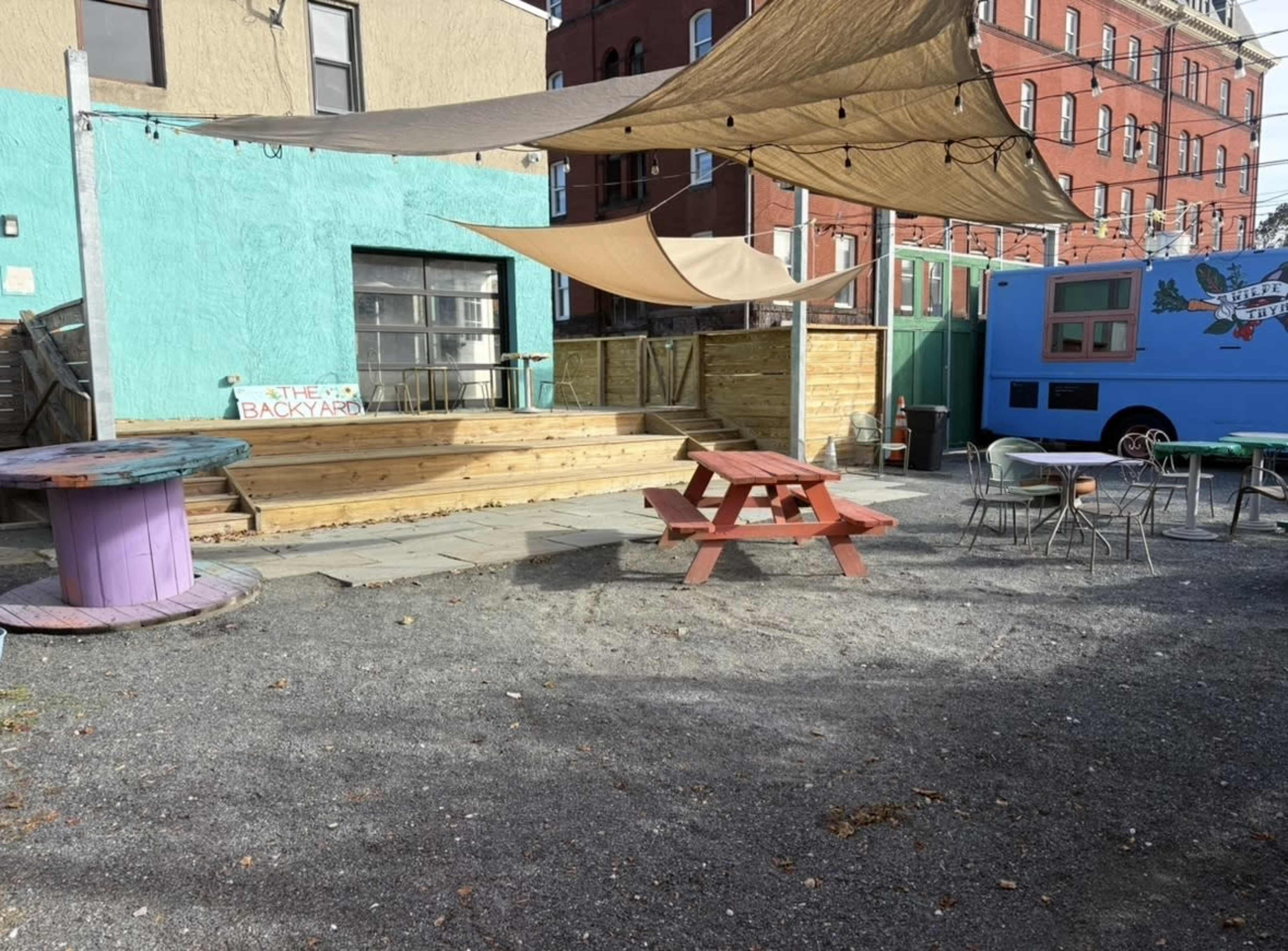 The image shows an outdoor space featuring a wooden deck, several tables, and large fabric canopies, set against a colorful building and a food truck.
