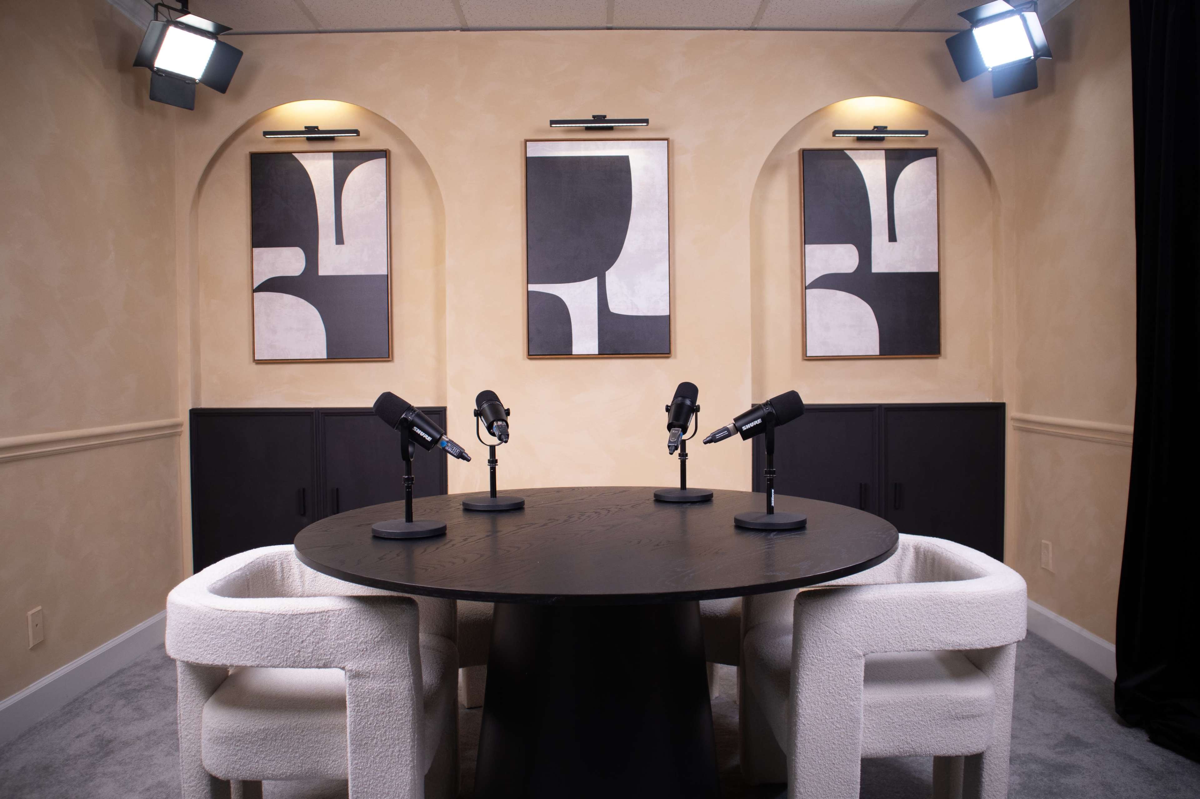 A neatly arranged interview setup with a round table at the center, three microphones, and three white chairs, alongside two abstract art pieces on the walls.