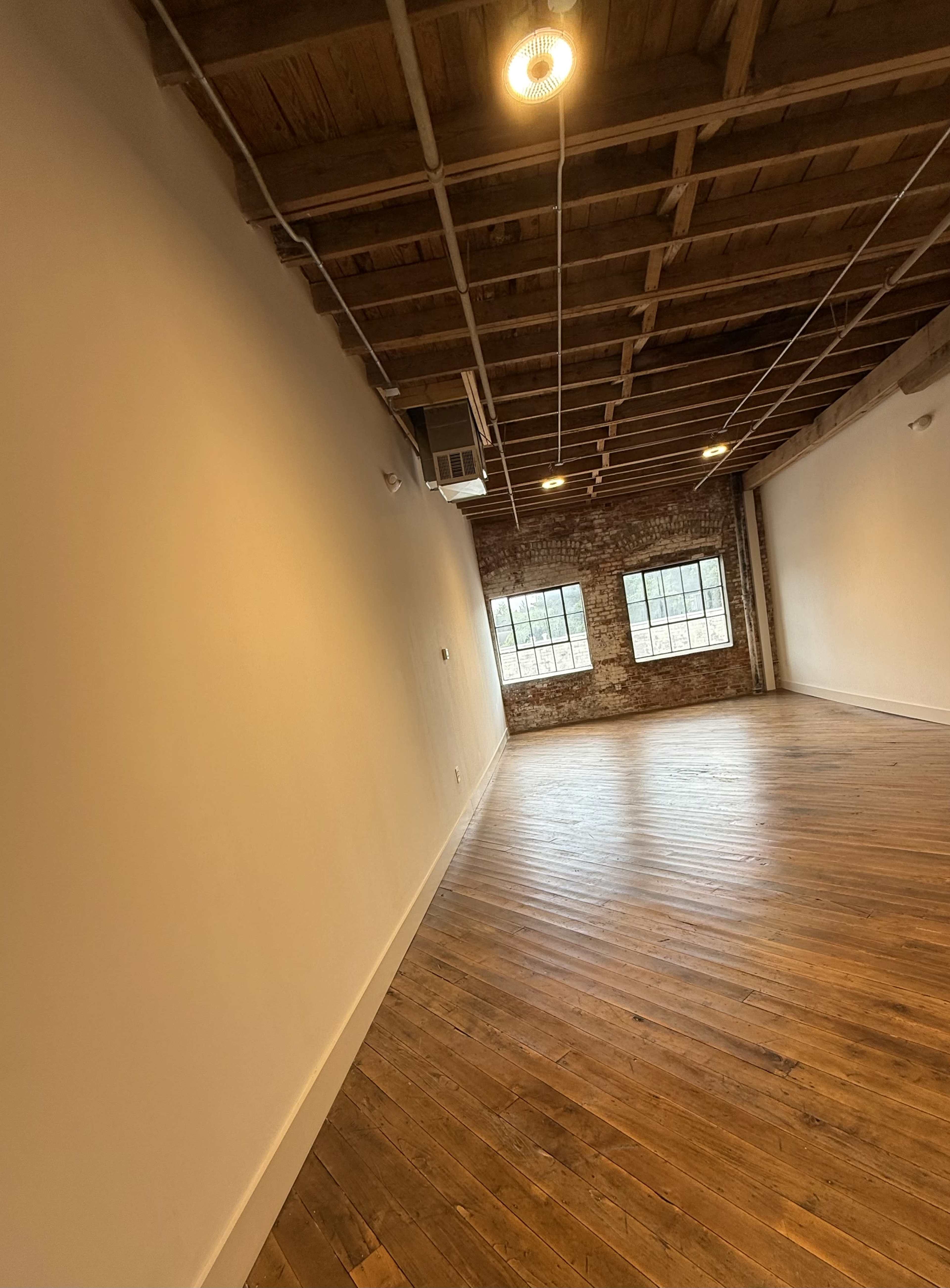 The image shows a spacious, empty room with hardwood floors, exposed brick walls, and large windows allowing natural light to enter.