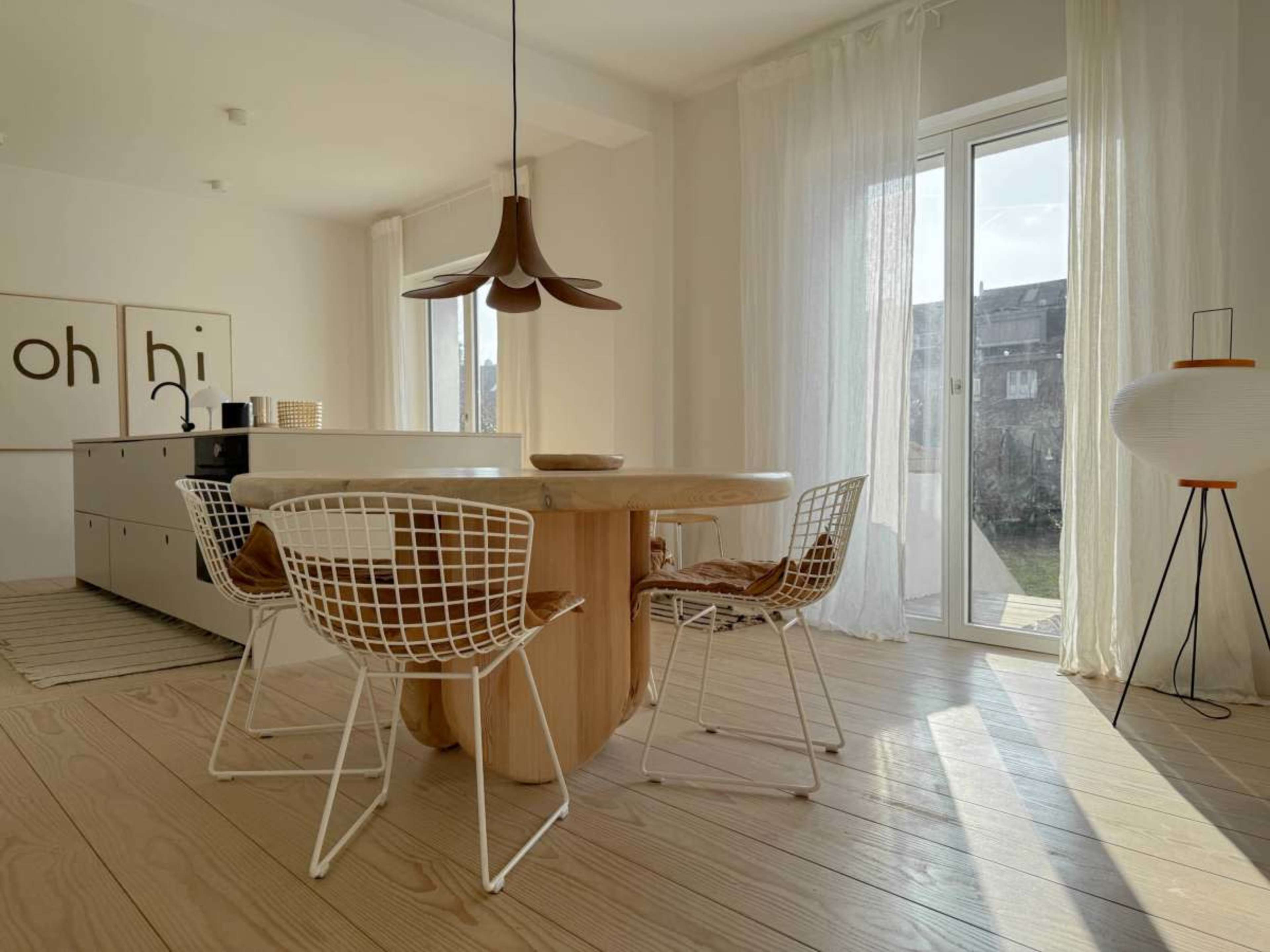 A modern dining area features a round wooden table surrounded by white wire chairs, with a large window letting in natural light.