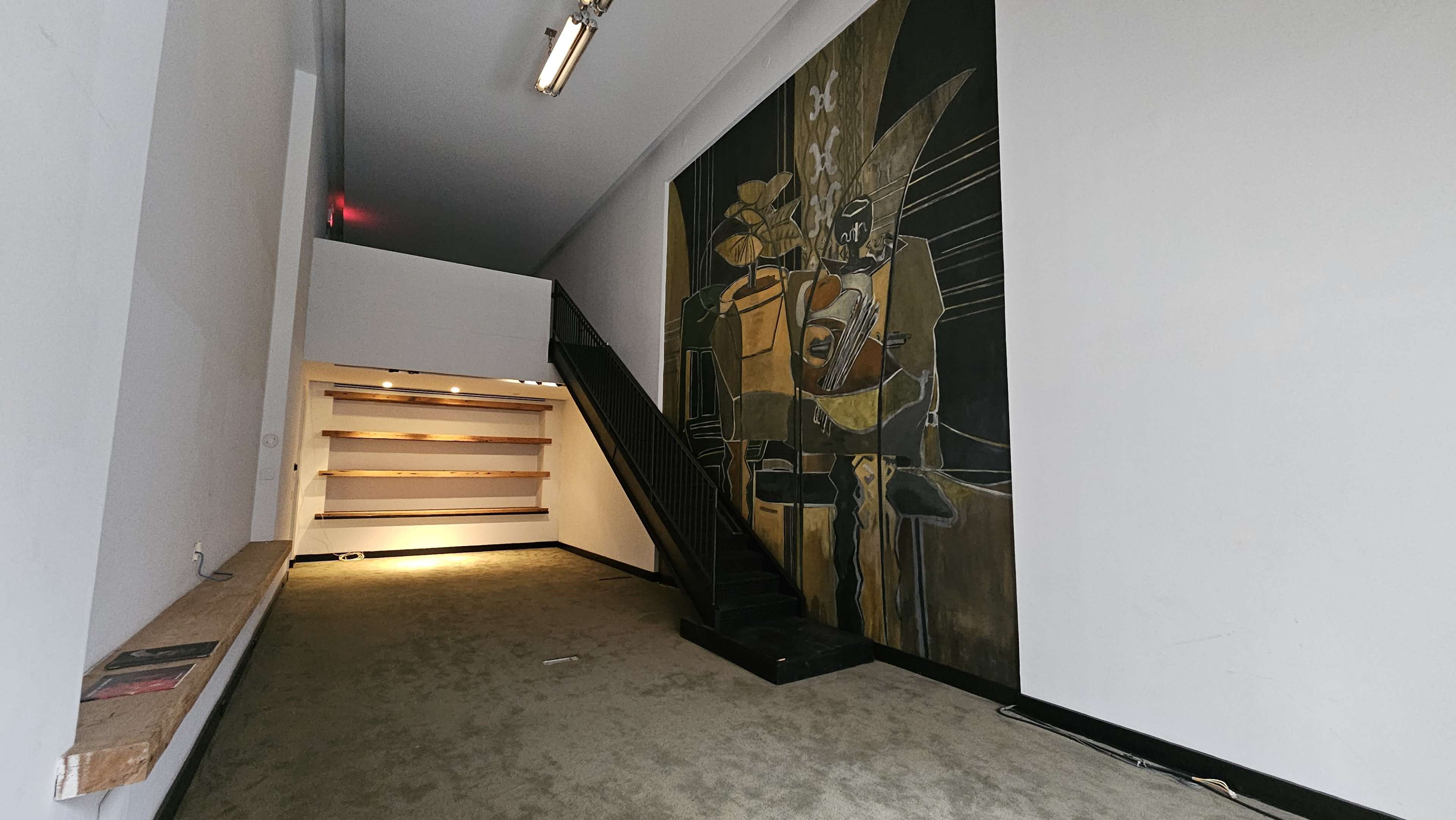 The image depicts a modern interior space featuring a large mural on one wall, a staircase to the left, and shelves along the opposite wall.