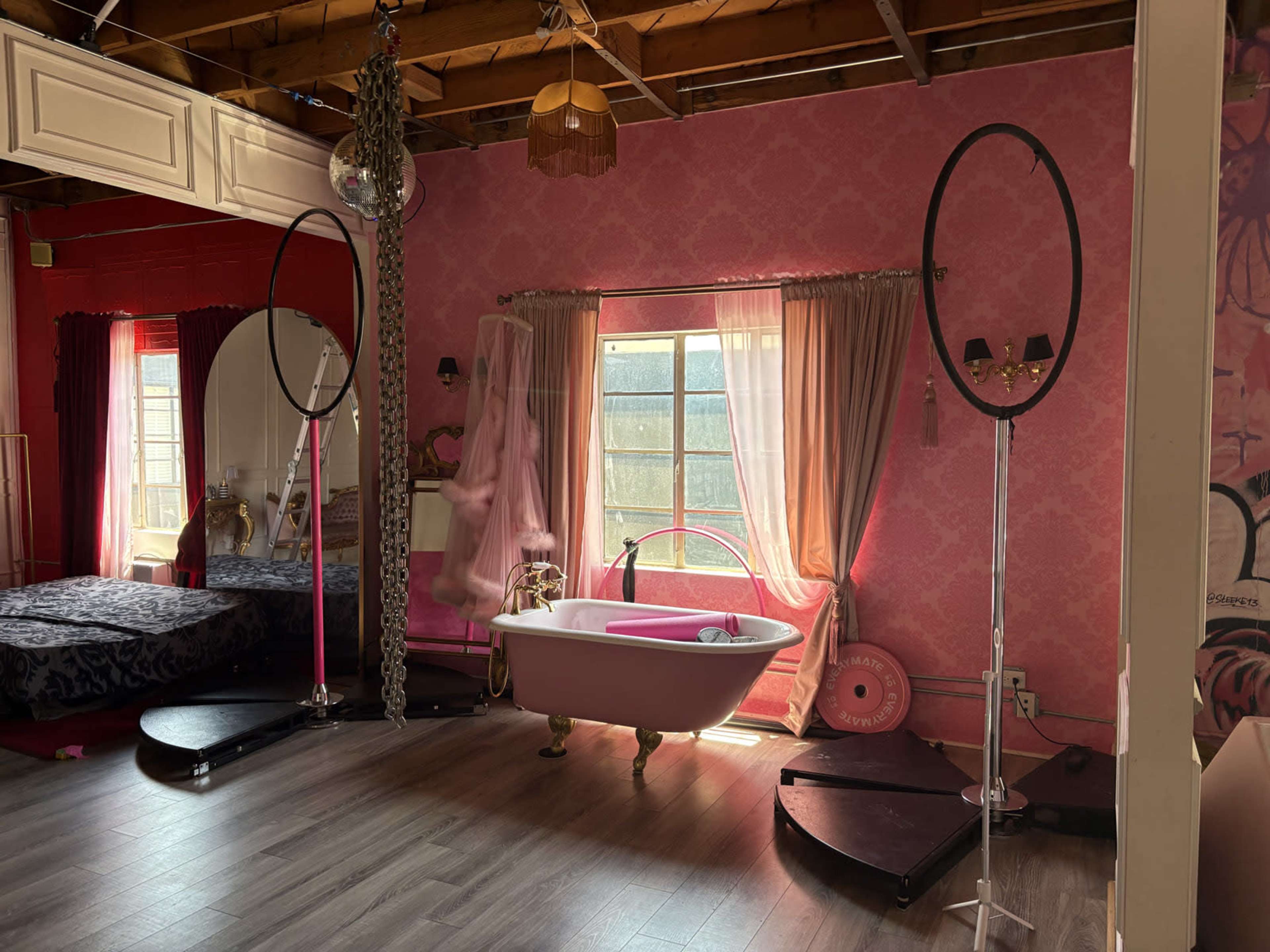 The image shows a room with a pink-themed decor featuring a vintage bathtub, a bed, and large windows with flowing curtains.