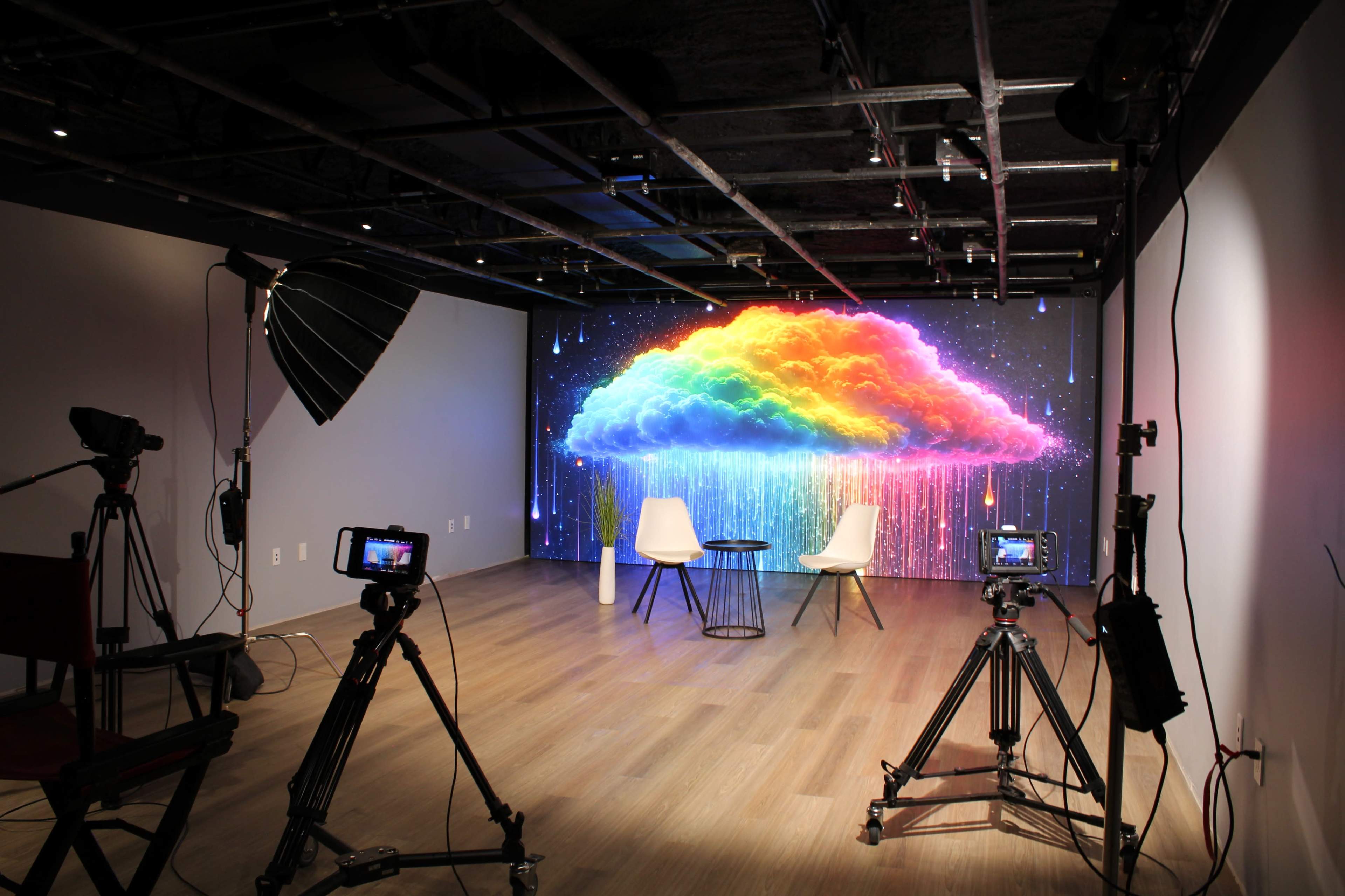 A modern studio setup features two chairs and a table in front of a vibrant, multicolored cloud mural, with cameras and lighting equipment positioned around the space.