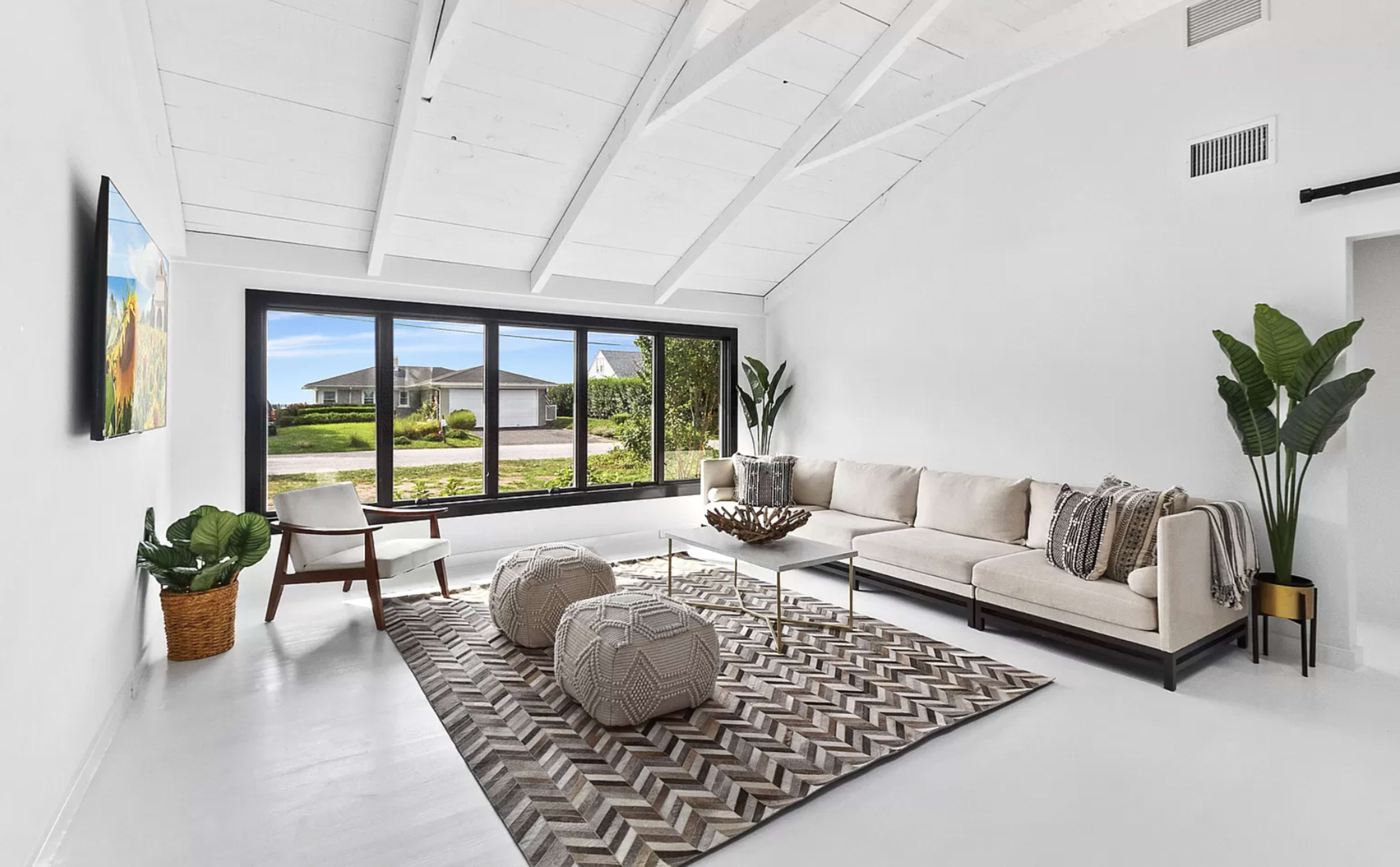 A bright, modern living room with a large window illuminating a sofa, coffee table, and two poufs on a patterned rug.