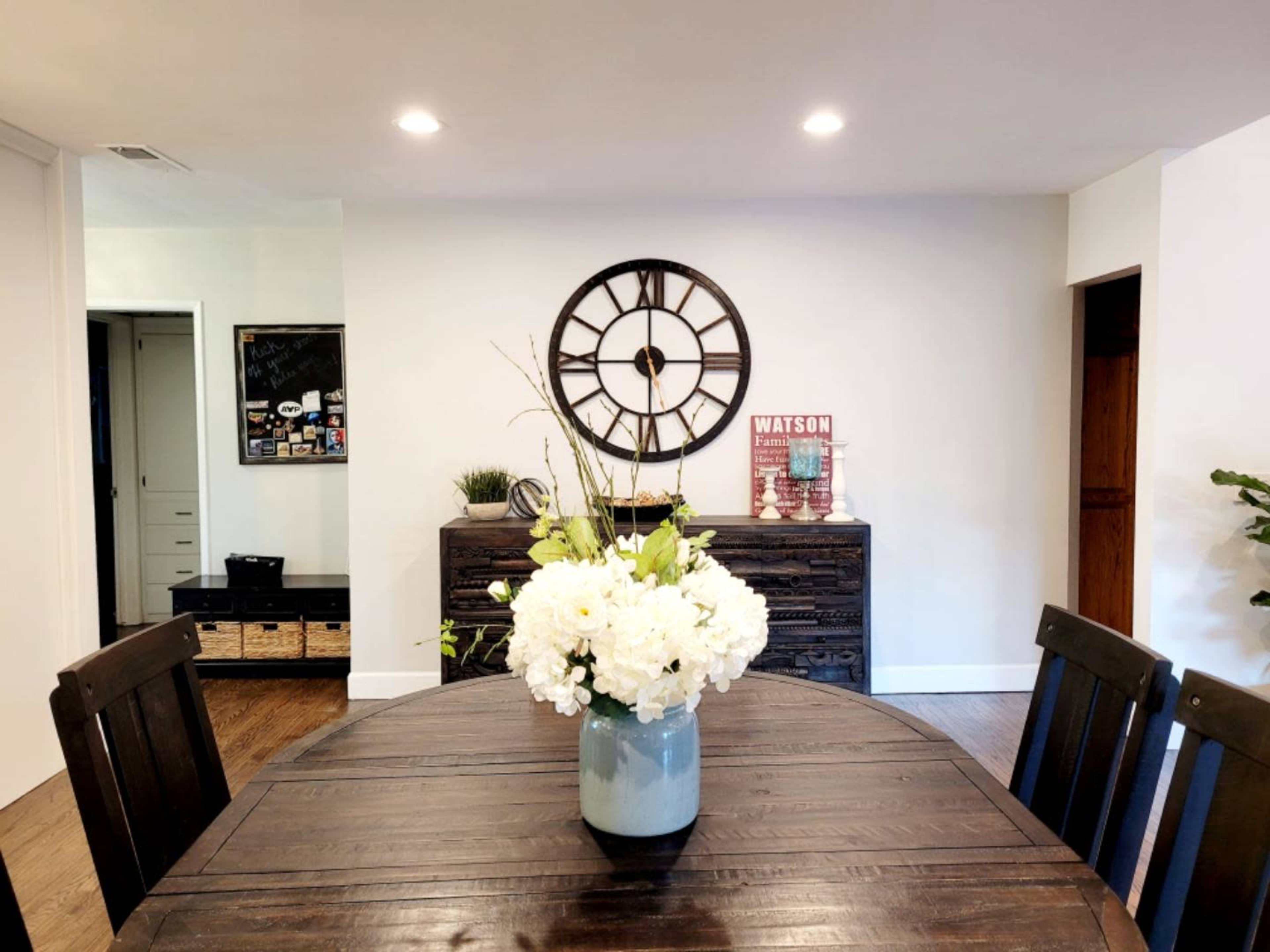 A round wooden dining table is set with a vase of white flowers, and a wall clock is prominently displayed behind it.