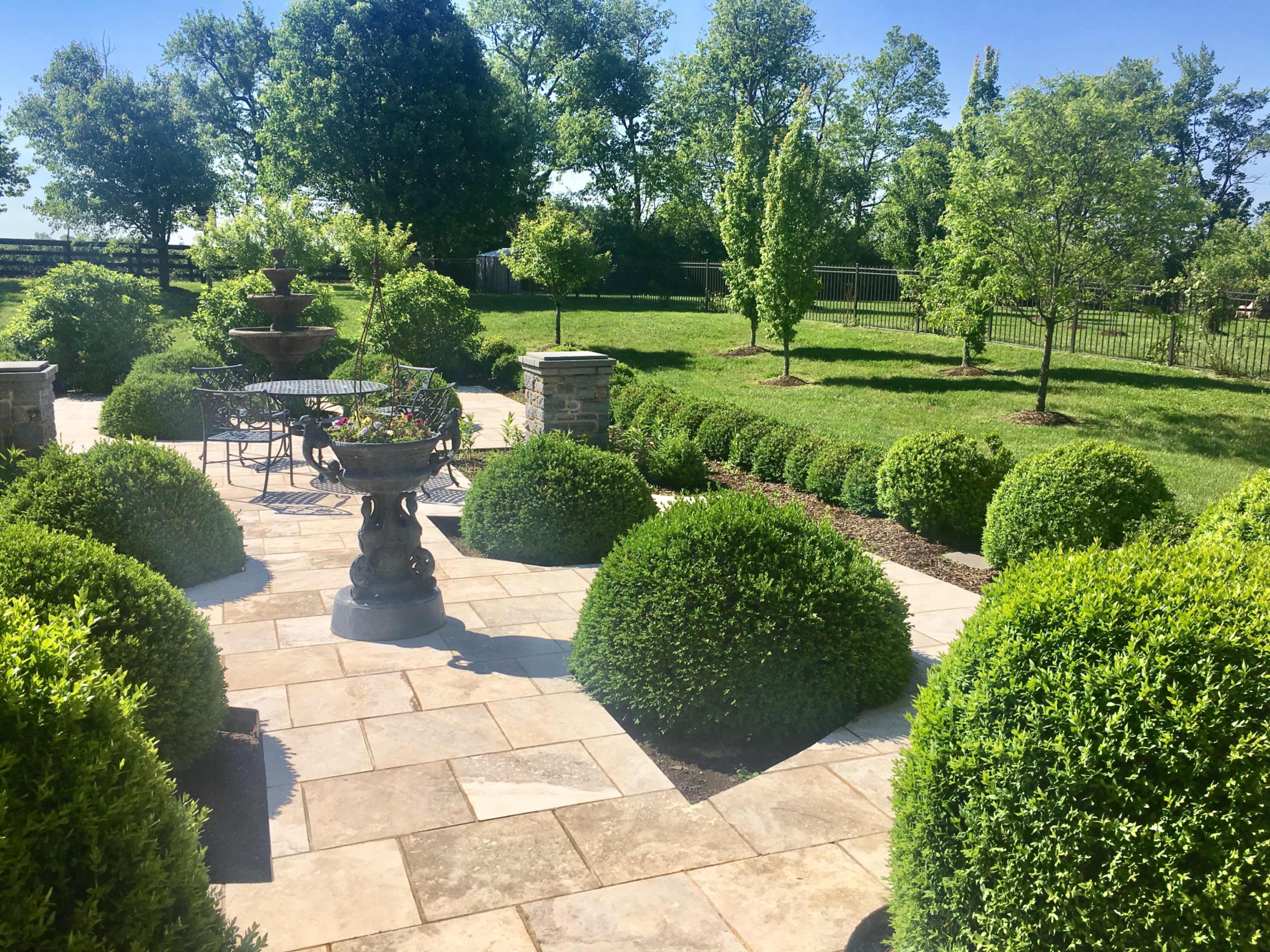 A well-maintained patio features circular hedges, a stone fountain, and a metal table with chairs, surrounded by a green lawn and trees.