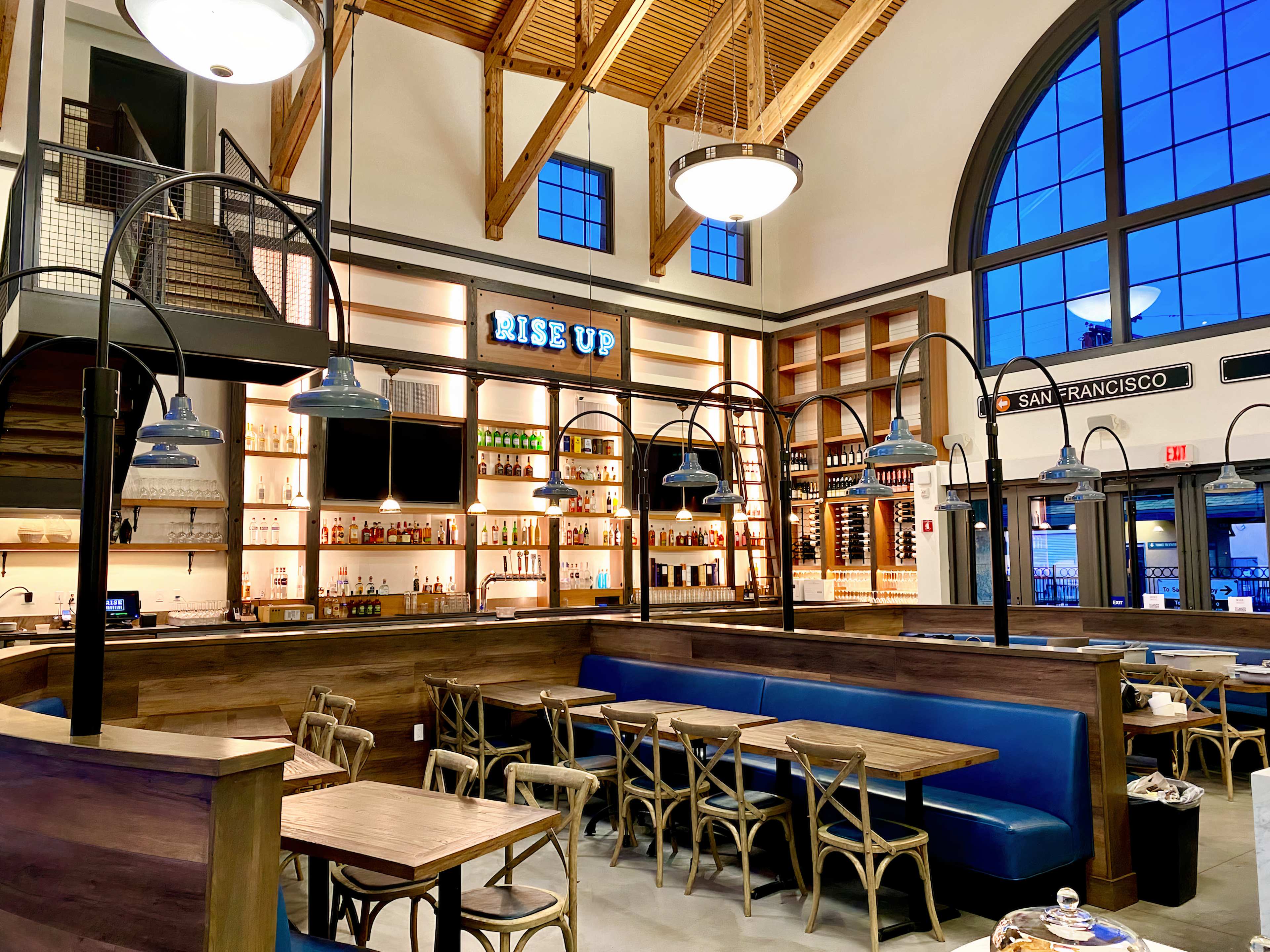 A modern restaurant interior with wooden furnishings, blue booth seating, and a bar displaying various bottles, all under a high ceiling with large windows.