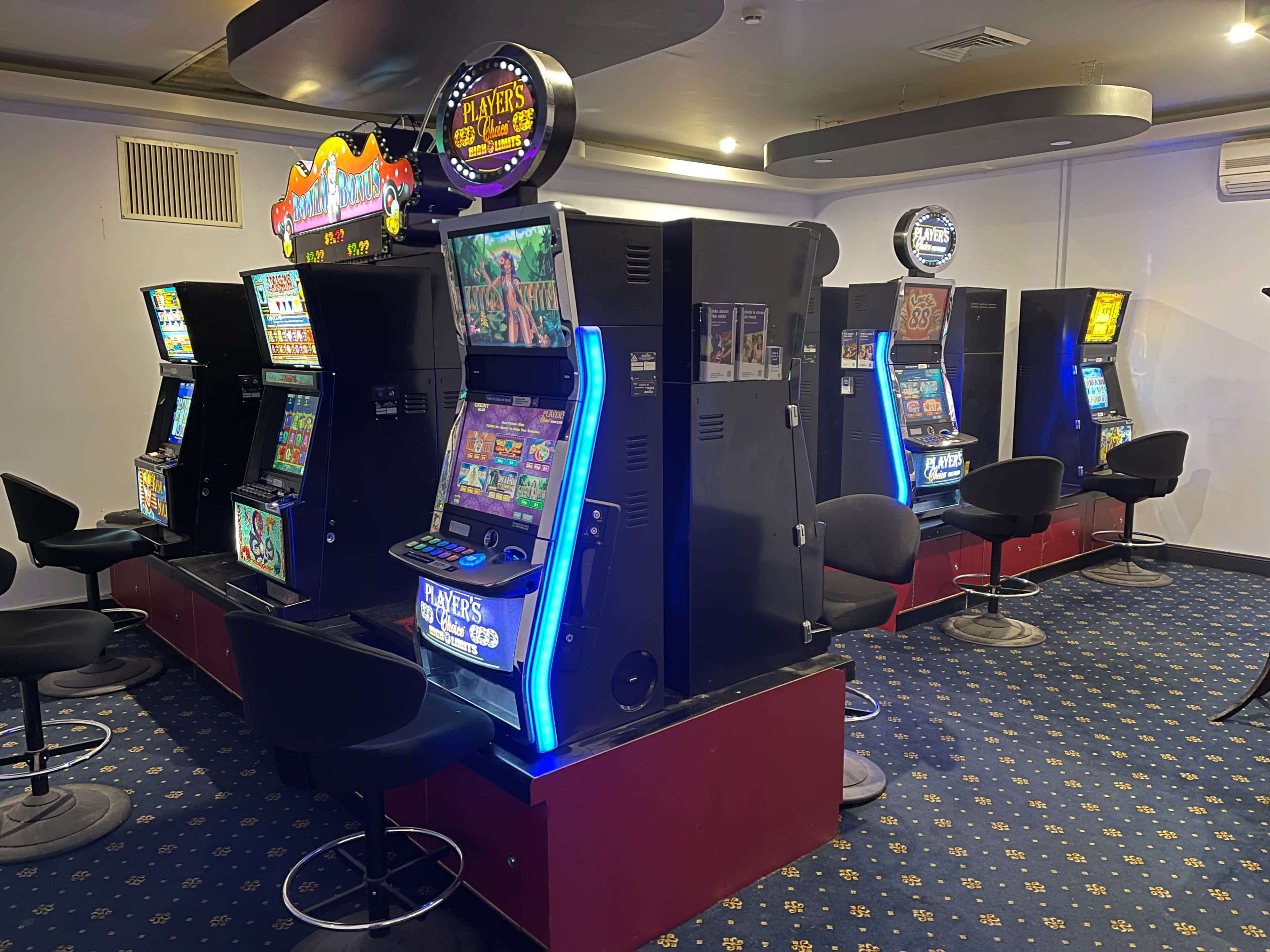 The image shows several video slot machines arranged in two rows, each with colorful screens, in a dimly lit room with patterned blue carpet.