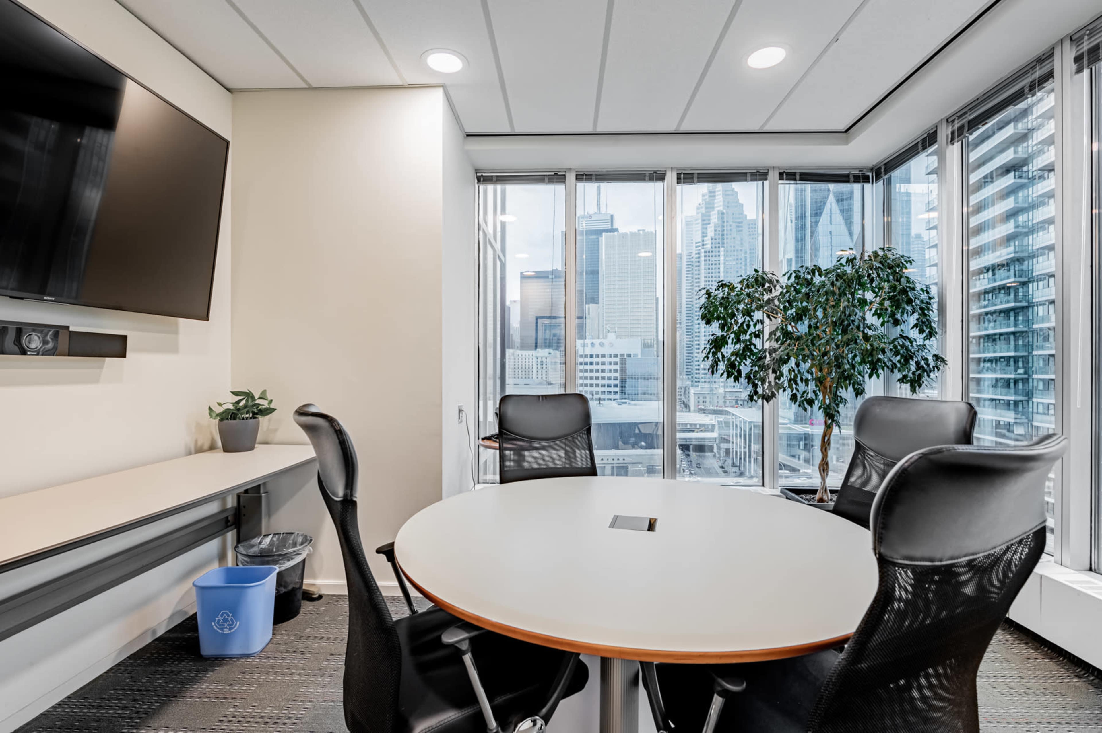 A modern conference room features a round table with four black chairs, a wall-mounted television, and large windows overlooking a city skyline.