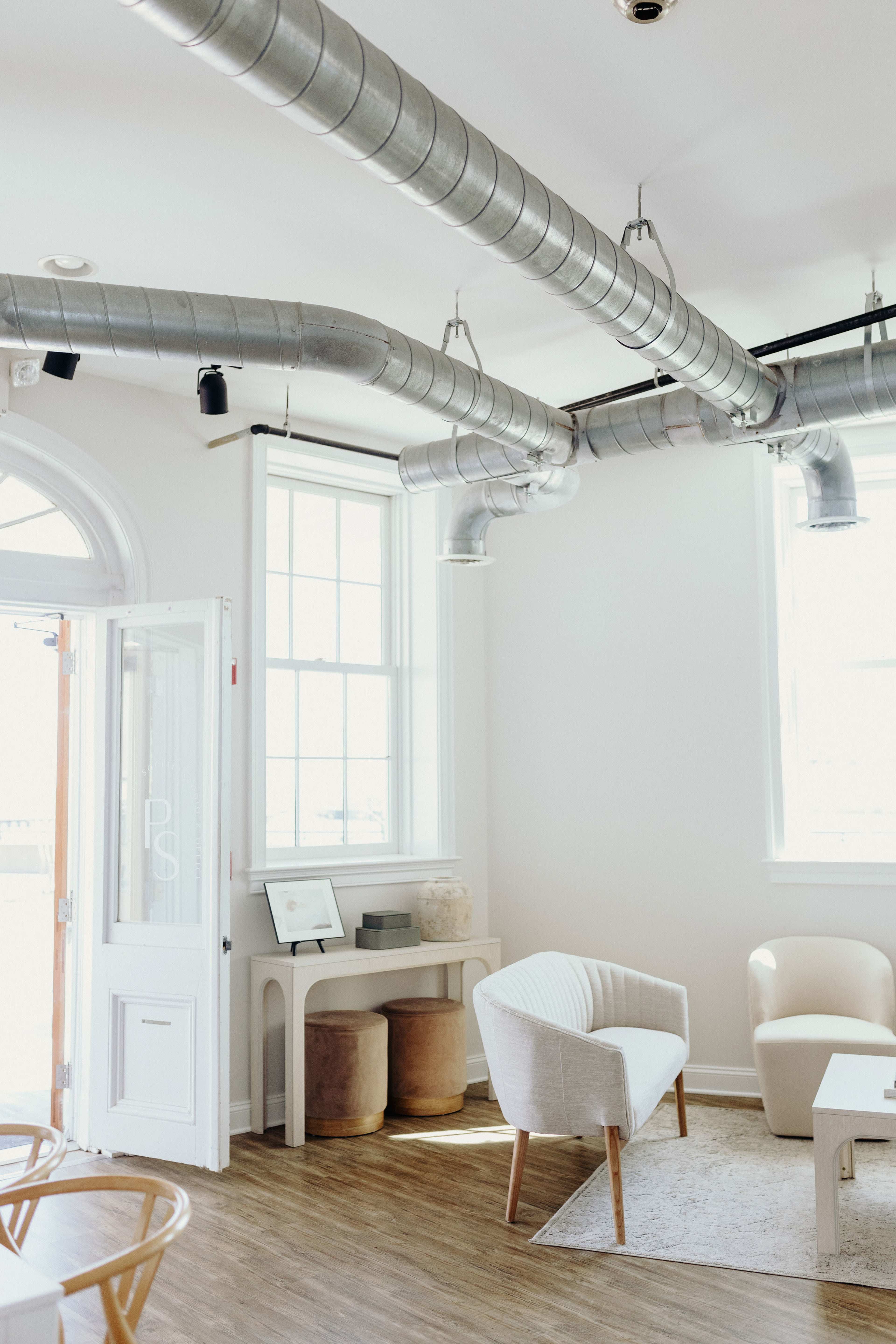 A bright, modern interior space featuring a white armchair, wooden flooring, and exposed ductwork along the ceiling.