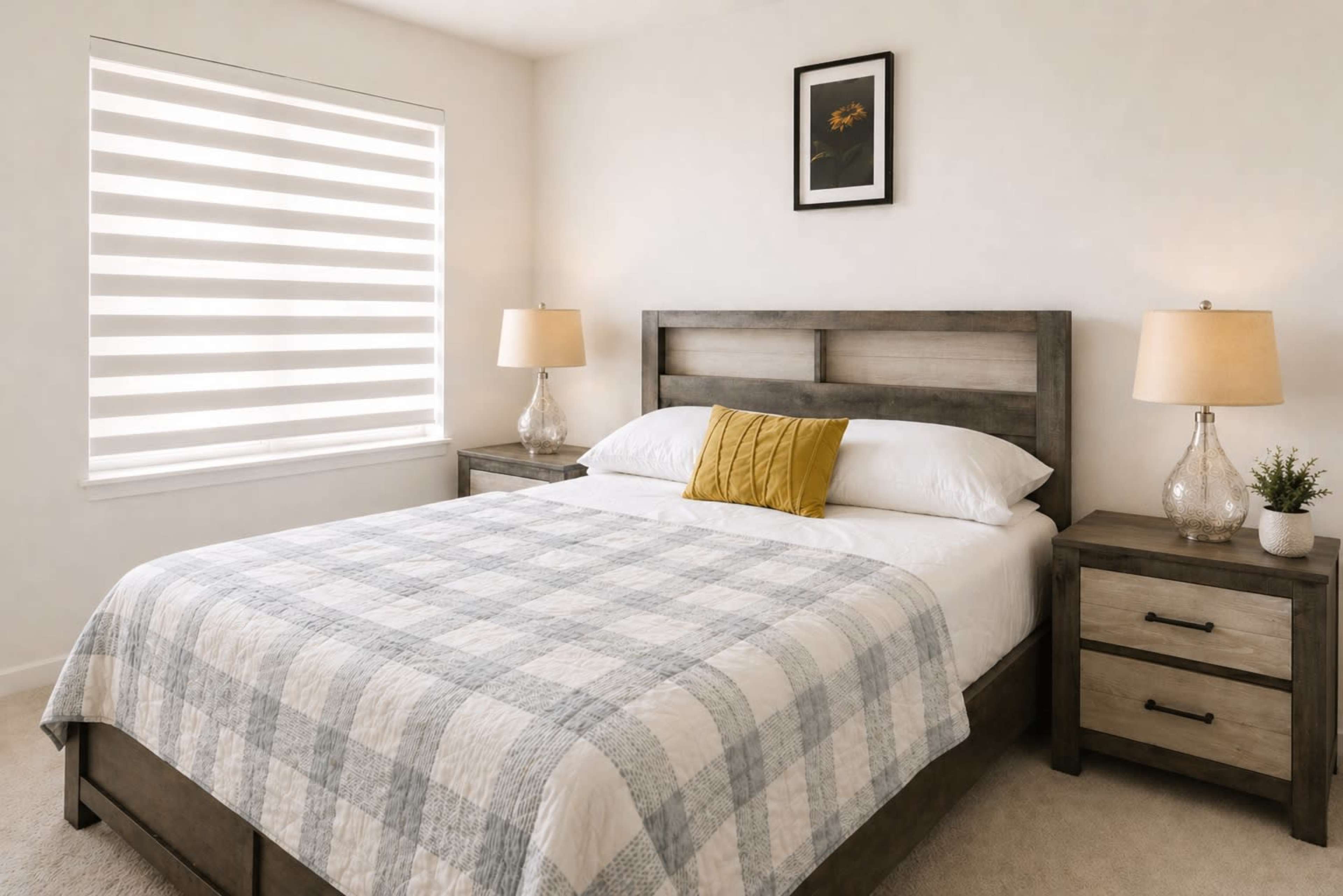 The image shows a well-lit bedroom featuring a bed with a plaid quilt, two bedside tables with lamps, and a framed picture on the wall.