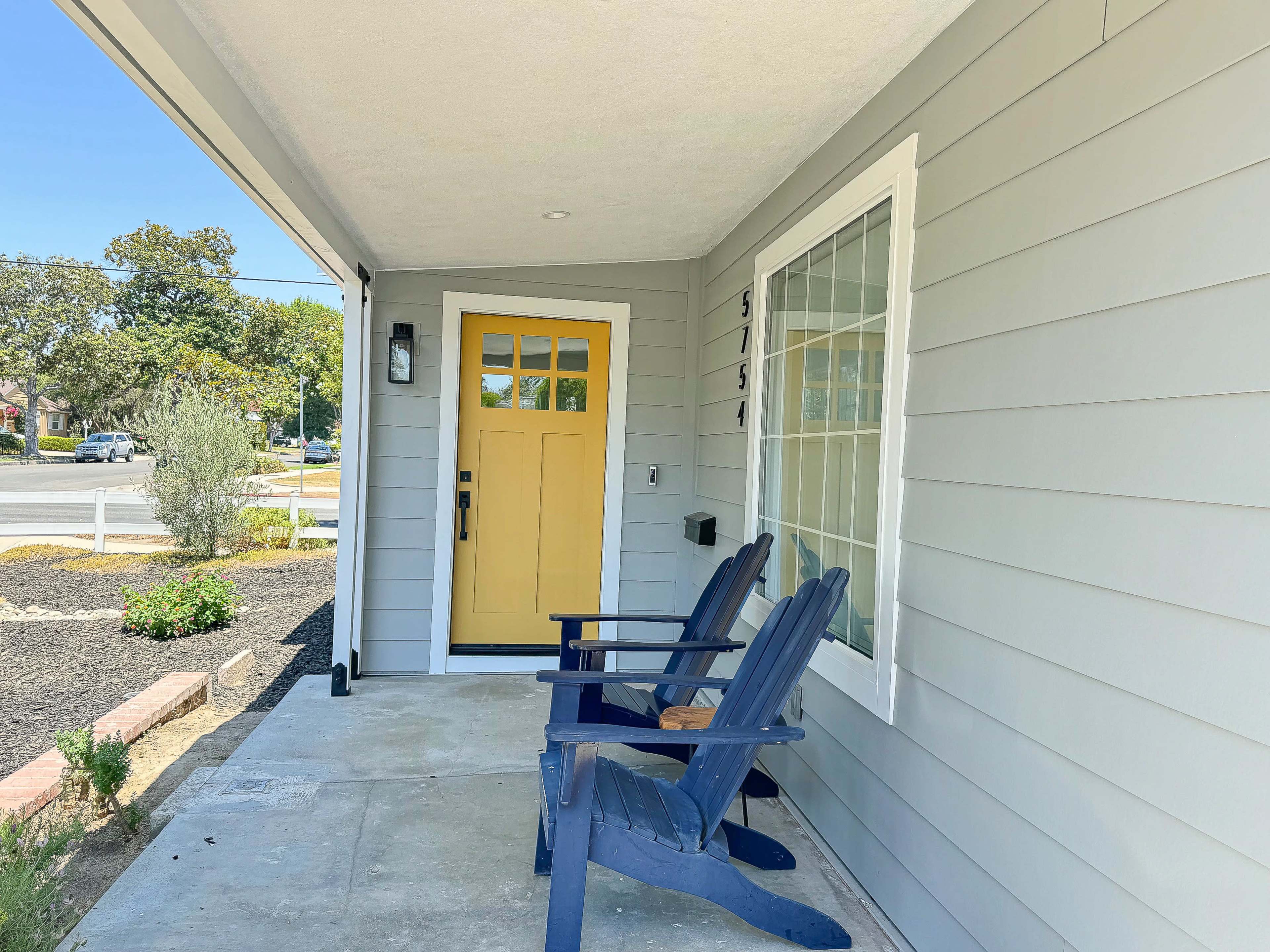 The image shows a home entrance featuring a yellow door, two blue adirondack chairs on a concrete porch, and surrounding plants.