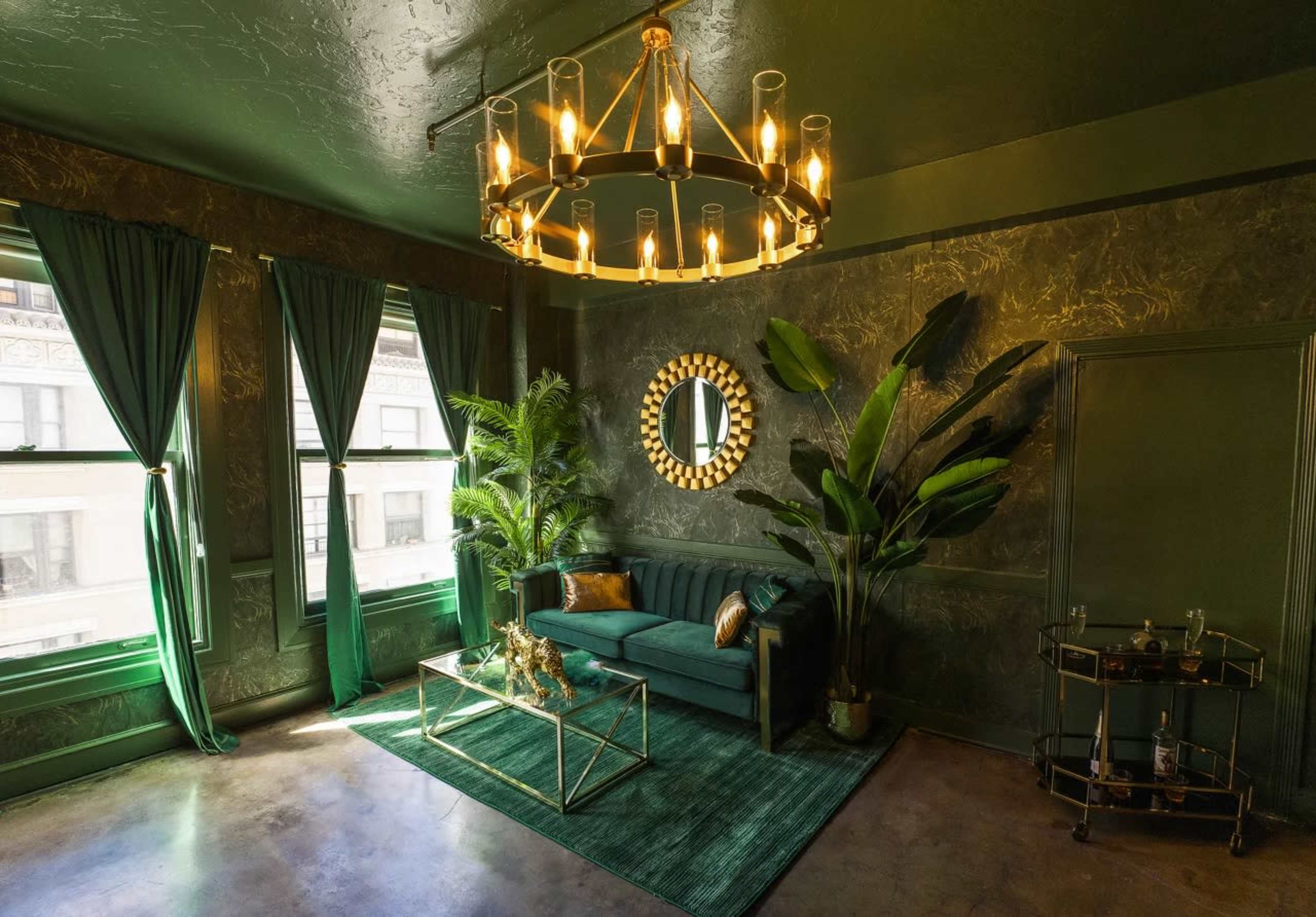 A stylish room features a green sofa and large plants, with a chandelier overhead and a glass coffee table in front of a mirror on the wall.