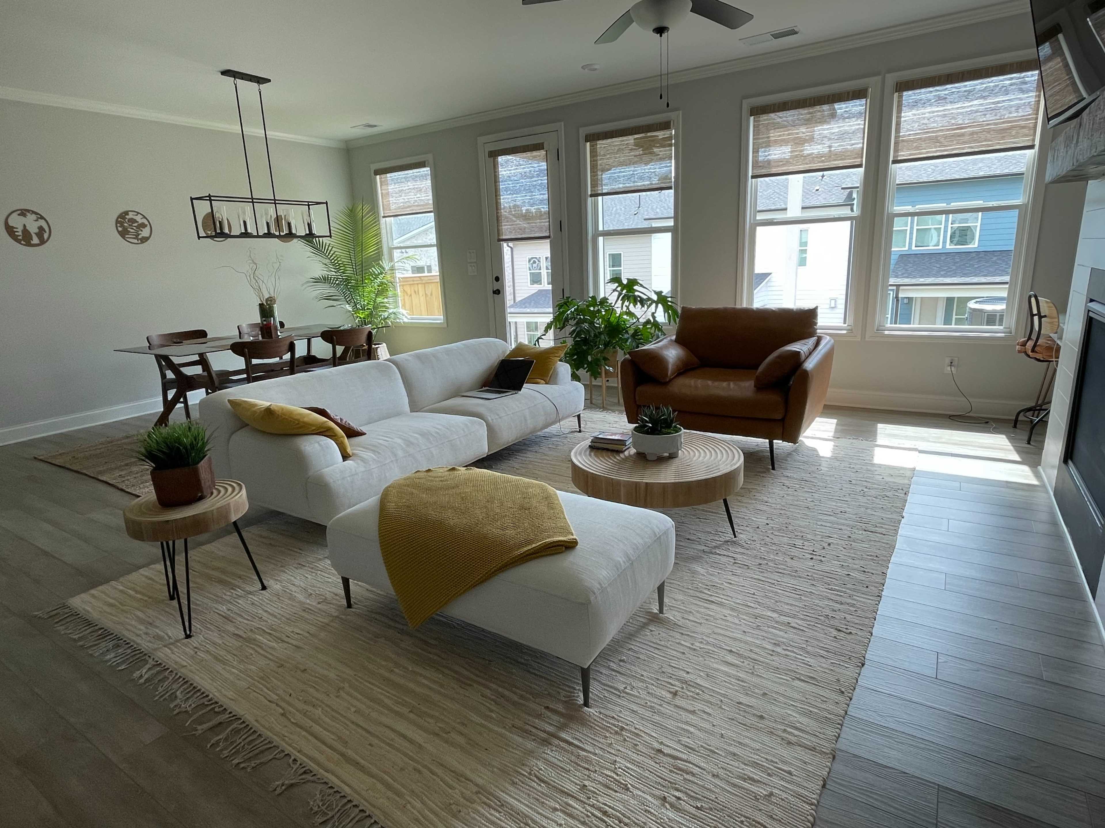 The image shows a modern living room featuring a large white sectional sofa, a brown leather armchair, a round wooden coffee table, and a dining area with a wooden table and chairs, adorned with plants and natural light from large windows.