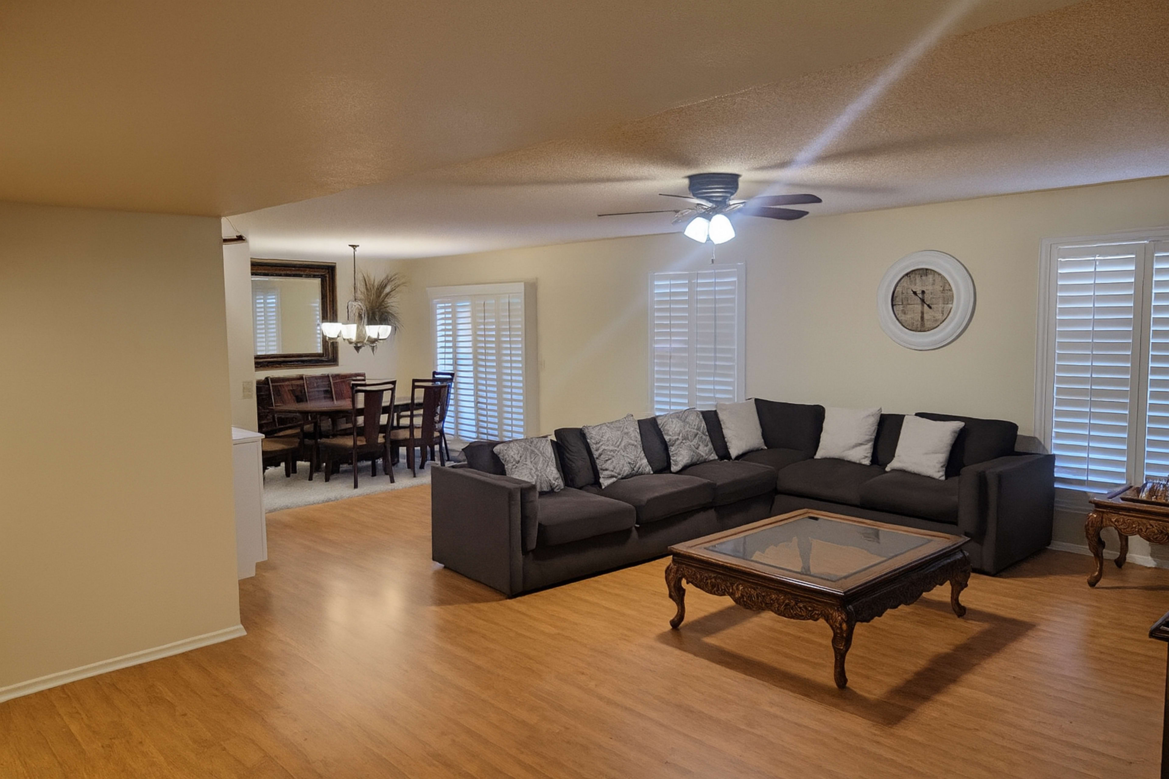 The image shows a spacious living area with a dark sectional sofa, a glass-topped coffee table, and a dining room visible in the background featuring a wooden dining table and chairs.