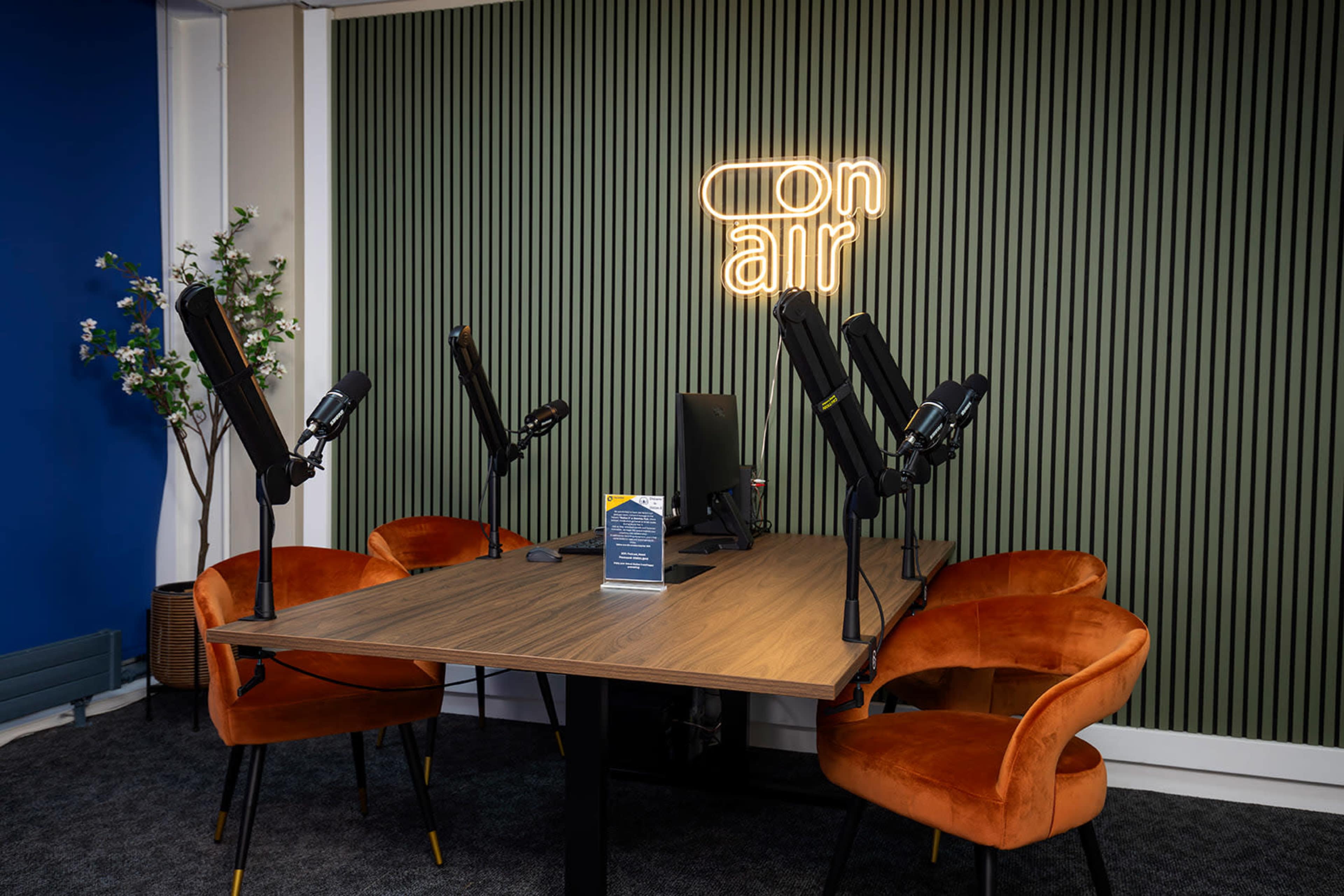 The image shows a podcast recording studio with a wooden table, four microphone setups, and two orange chairs, against a green paneled wall with a neon sign that reads "on air."
